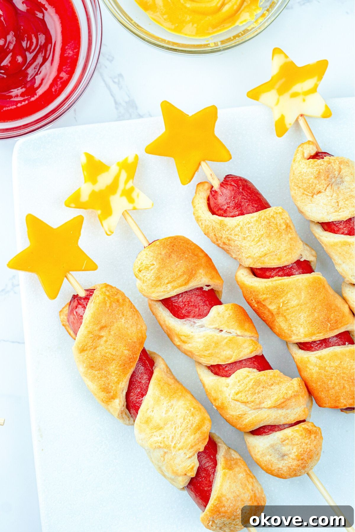 Dough wrapped hot dogs with cheese stars stuck into skewers, served with ketchup and mustard.