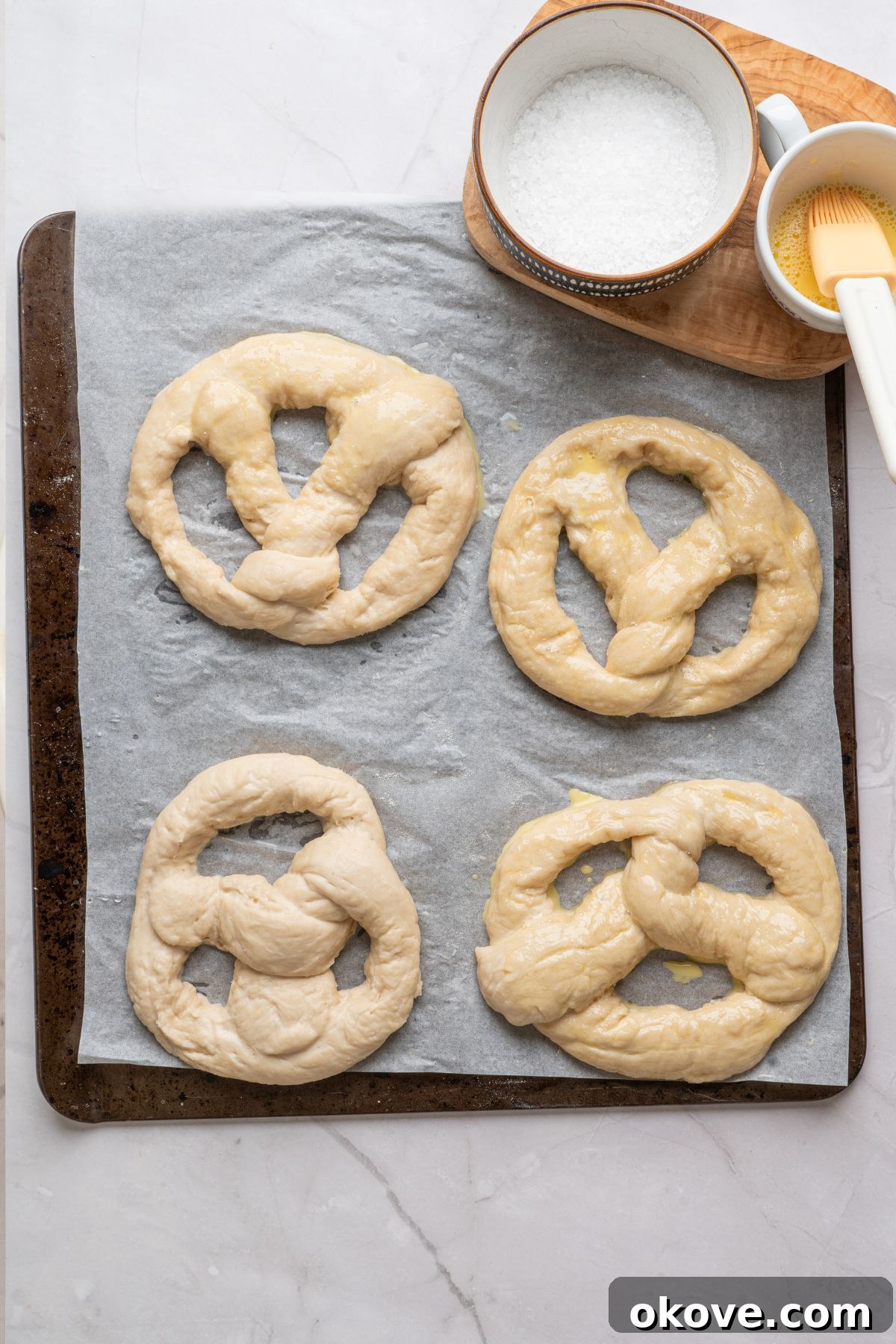 Pillowy Soft Pretzels 11 Boiled pretzels being brushed with egg wash and salt before baking, on a parchment paper lined baking sheet.