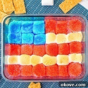 Red white and blue marshmallows over chocolate in a clear baking dish with graham crackers on the table for dipping.