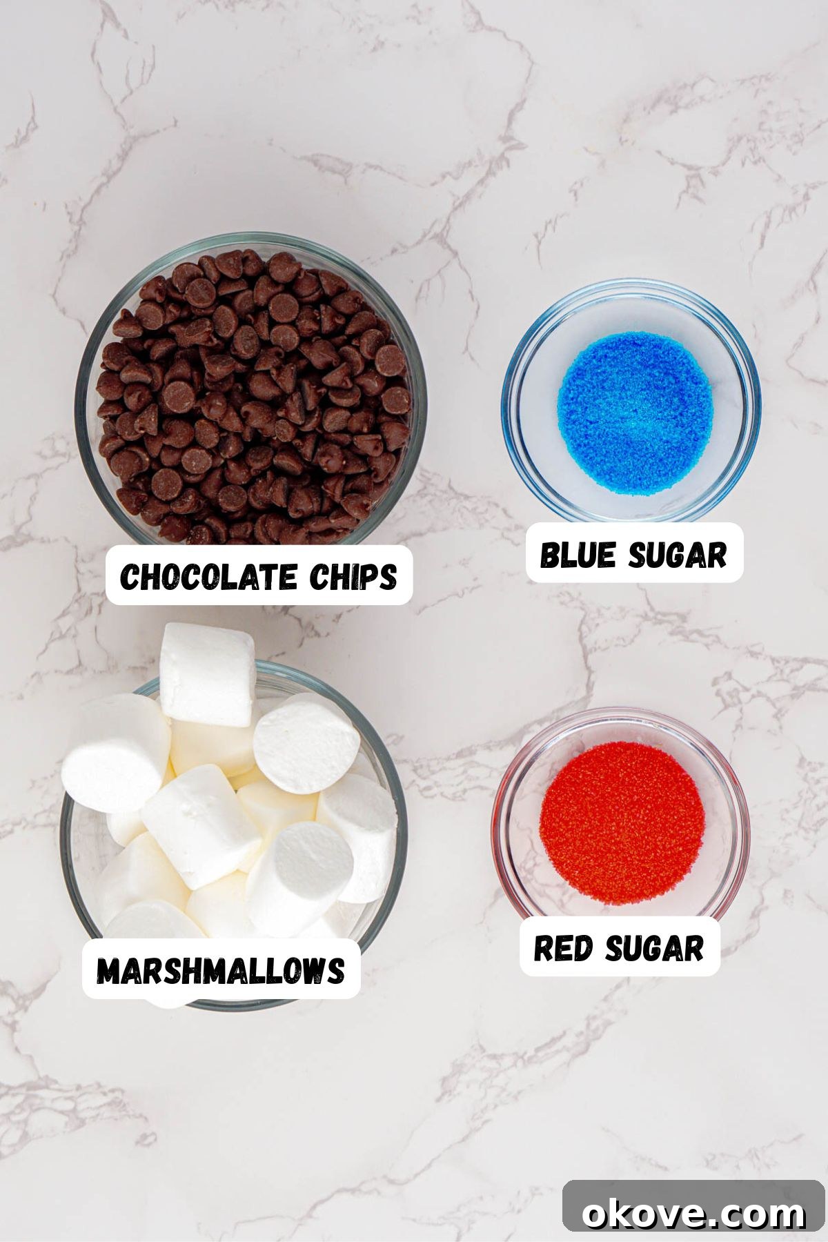 Red, White, and Gooey S'mores Dip 3 Small glass jars containing marshmallows, chocolate chips, red sanding sugar, and blue sanding sugar, arranged on a marble tabletop.
