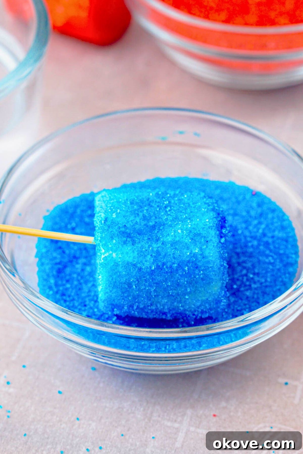 Red, White, and Gooey S'mores Dip 5 A marshmallow on a toothpick being dipped into a bowl of blue sanding sugar, creating a vibrant blue coating.