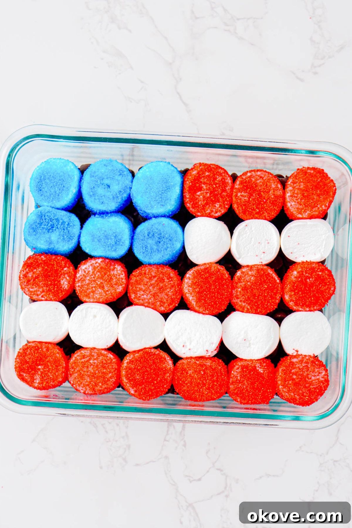 Red, White, and Gooey S'mores Dip 8 A clear glass baking dish with a layer of chocolate chips on the bottom, meticulously topped with red, white, and blue marshmallow halves arranged in a flag pattern.