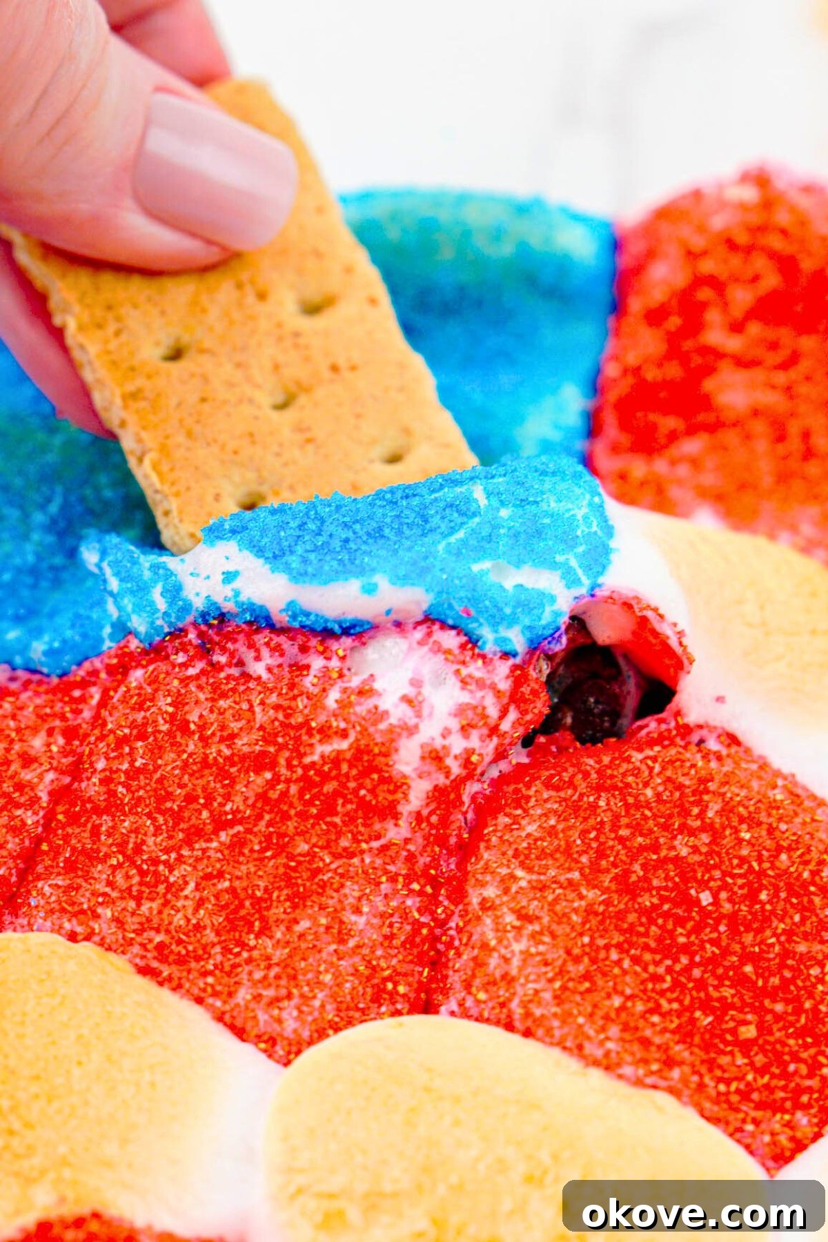 Red, White, and Gooey S'mores Dip 9 A graham cracker being dipped into the freshly baked and melted Patriotic S'mores Dip, showing gooey chocolate and toasted marshmallows.