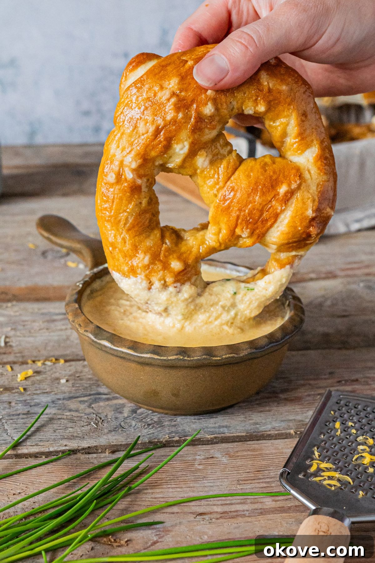 A freshly baked soft pretzel being dipped into a rustic bowl of rich, creamy beer cheese dip, beautifully garnished with fresh green herbs.