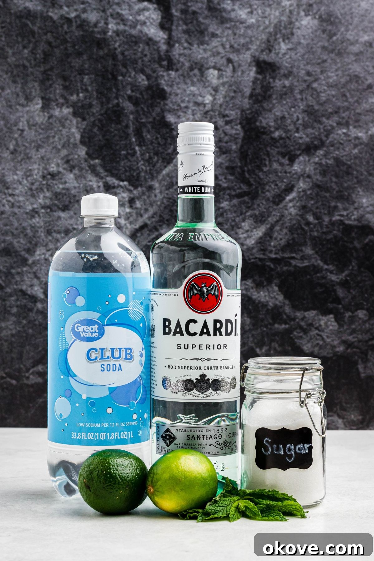 Bottles of white rum and sparkling club soda, alongside a jar of sugar, fresh limes, and vibrant mint leaves, all arranged on a elegant marble table.