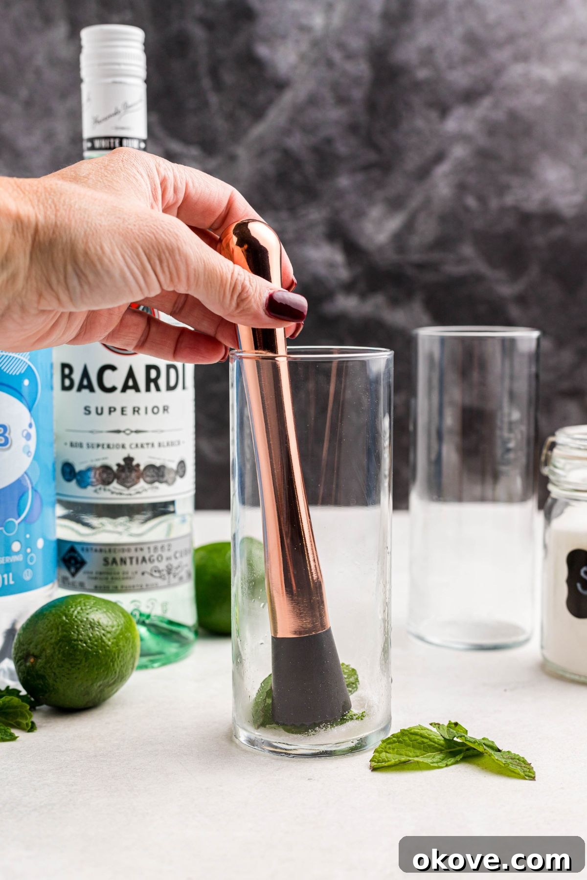 Sugar, fresh lime juice, and vibrant mint leaves being gently muddled together in a clear highball glass.
