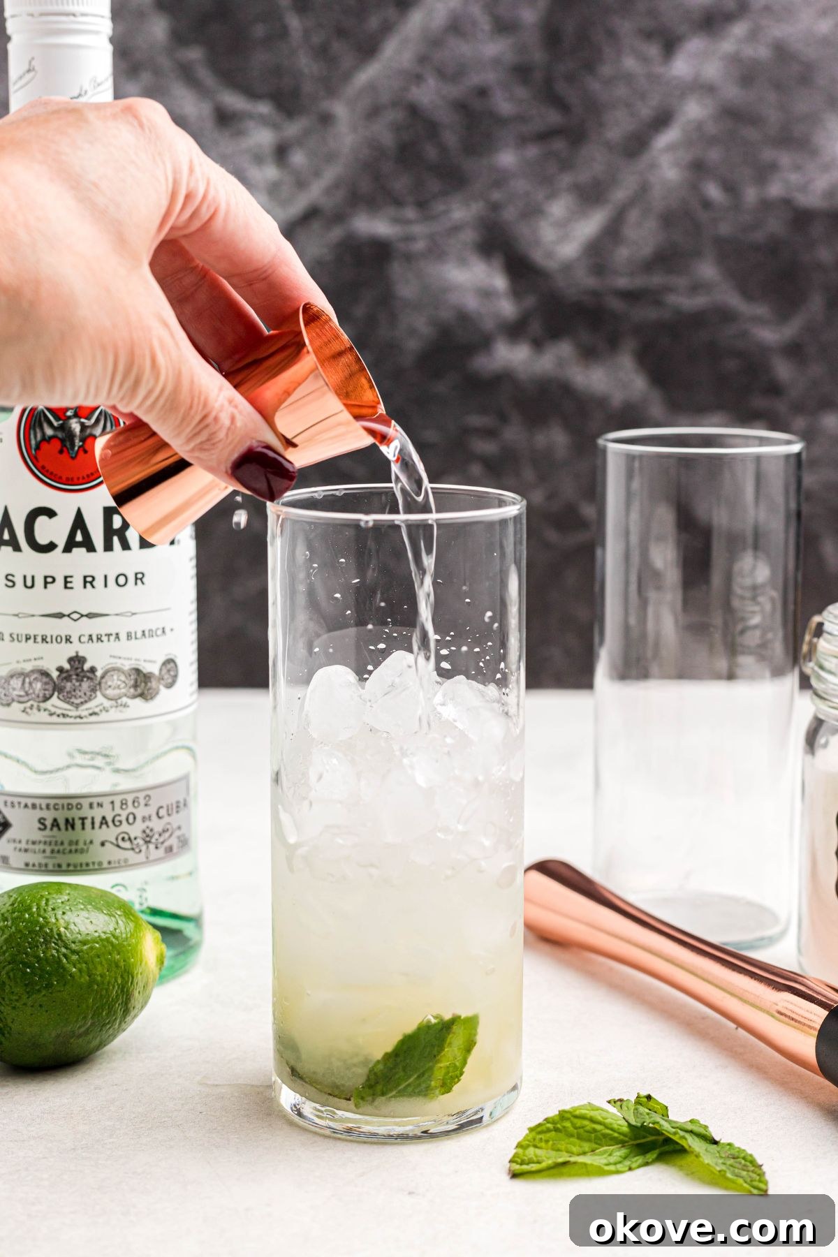 White rum being poured into a tall clear glass already containing ice, muddled mint leaves, sugar, and lime juice.