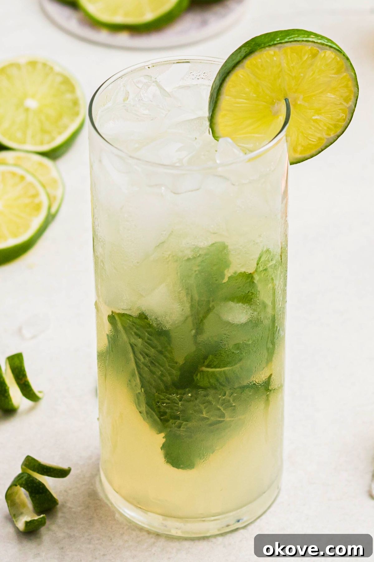 Bright yellow mojito drink in a tall chilled glass, featuring two green striped straws, with limes and lime curls on the marble table.