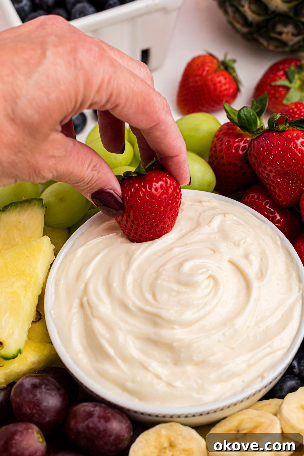 Sweet and Creamy Fruit Dip 6 A hand gently dips a vibrant red strawberry into a bowl of creamy white fruit dip, surrounded by an abundant assortment of fresh, colorful fruits like grapes, bananas, and blueberries, all arranged on a white platter.