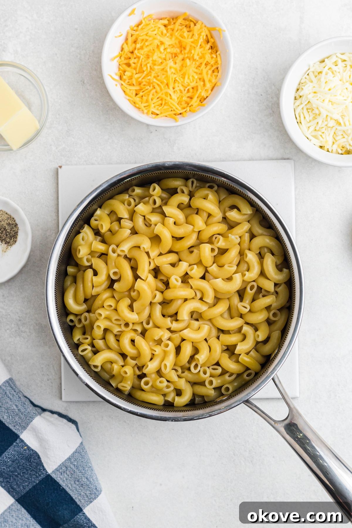 Golden Baked Mac 4 Lightly boiled elbow macaroni in a saucepan, surrounded by shredded cheese and seasonings.
