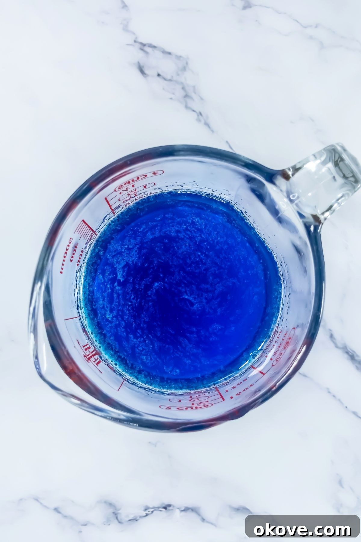 Tri-Color Celebration Shots 4 Blue Jello powder being mixed with hot water and vodka in a clear glass bowl on a marble table, preparing the first layer.