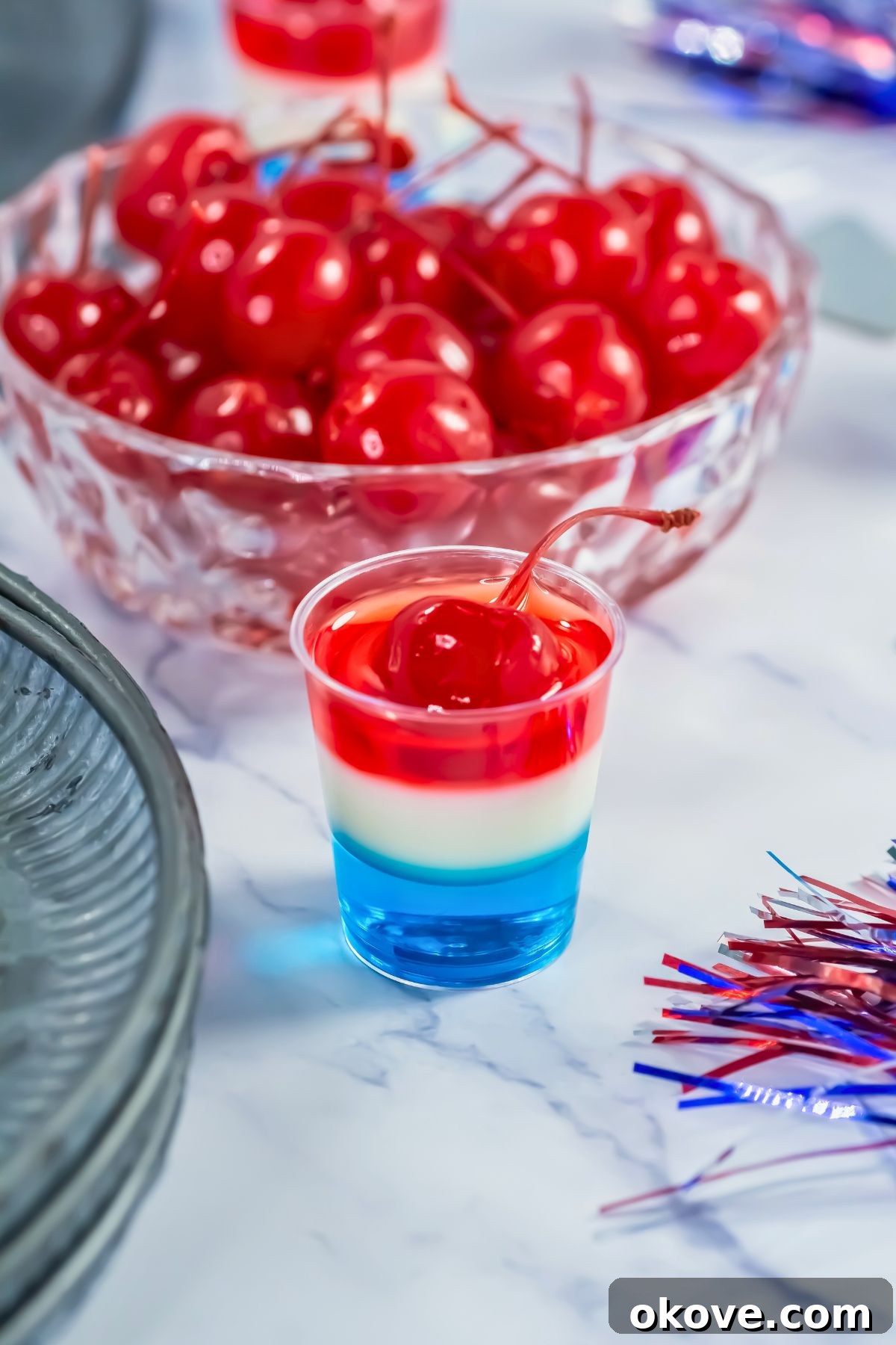 Tri-Color Celebration Shots 10 Perfectly layered red, white, and blue jello shot in a small glass on a marble table, with a bowl of red cherries in the background.