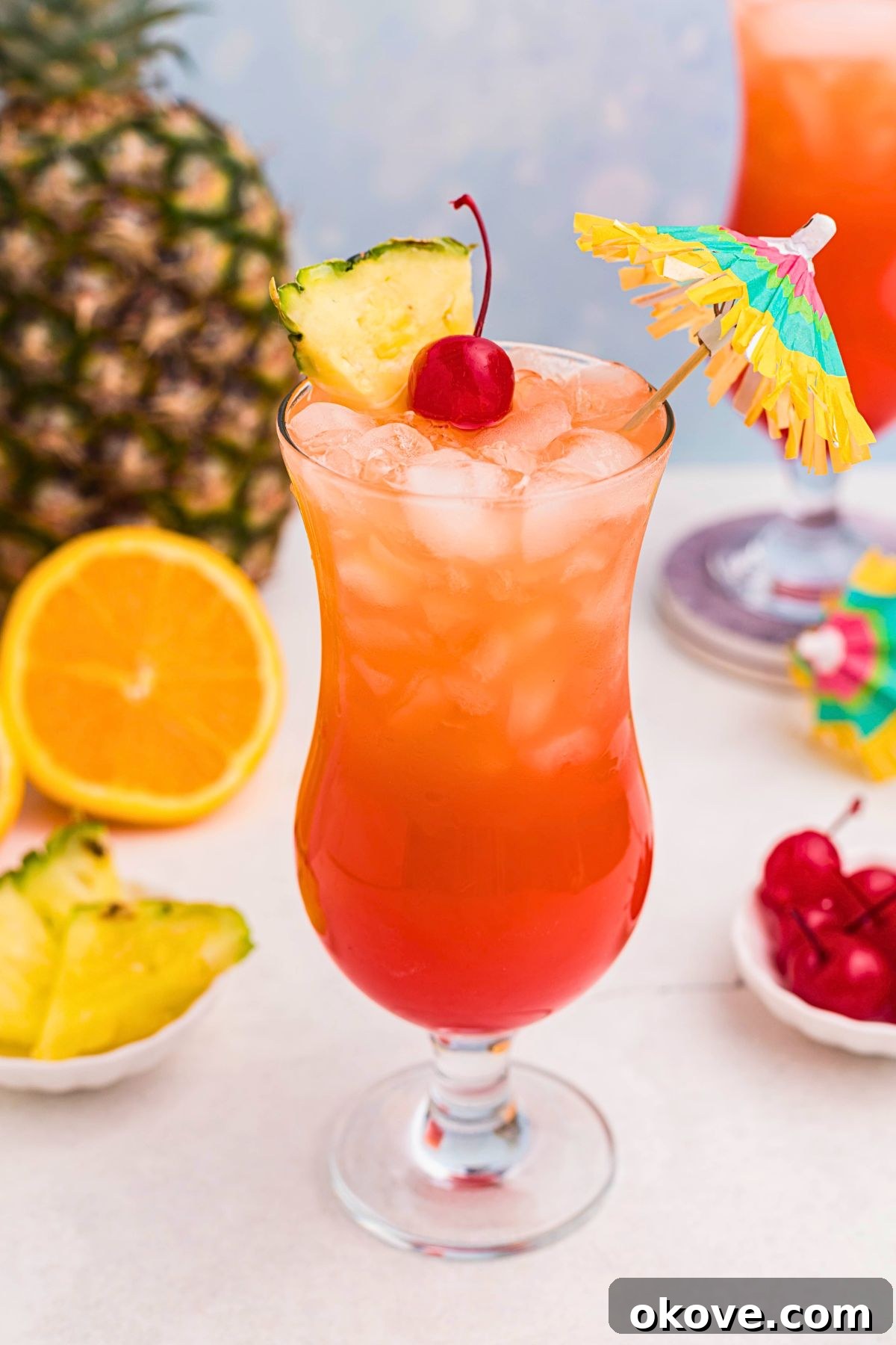 Red and orange drink mixed together, with a paper umbrella, in a hurricane glass with orange slices and cherries on the marble table.