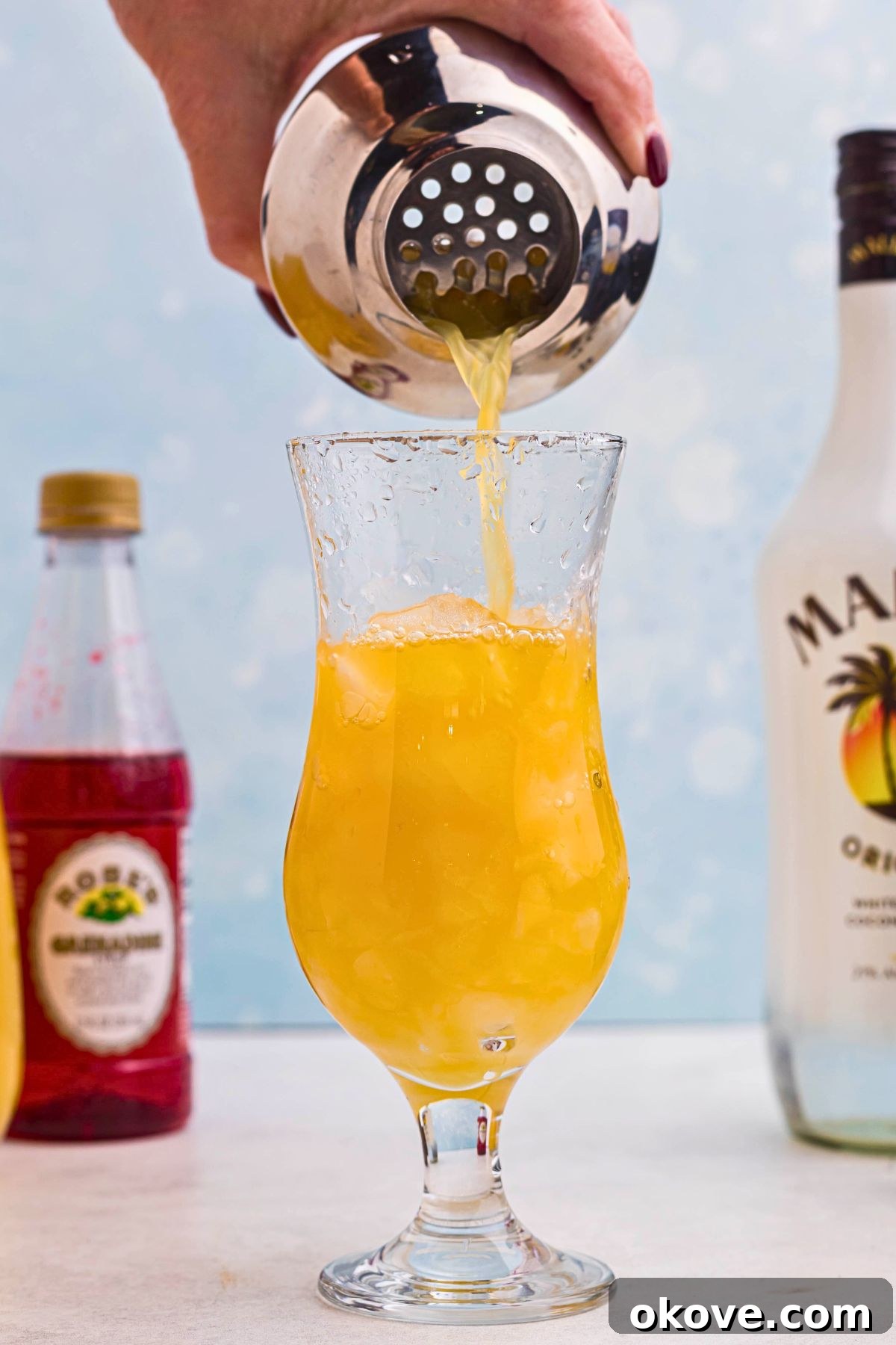 Orange drink being poured into hurricane glass over ice, from a cocktail shaker. 