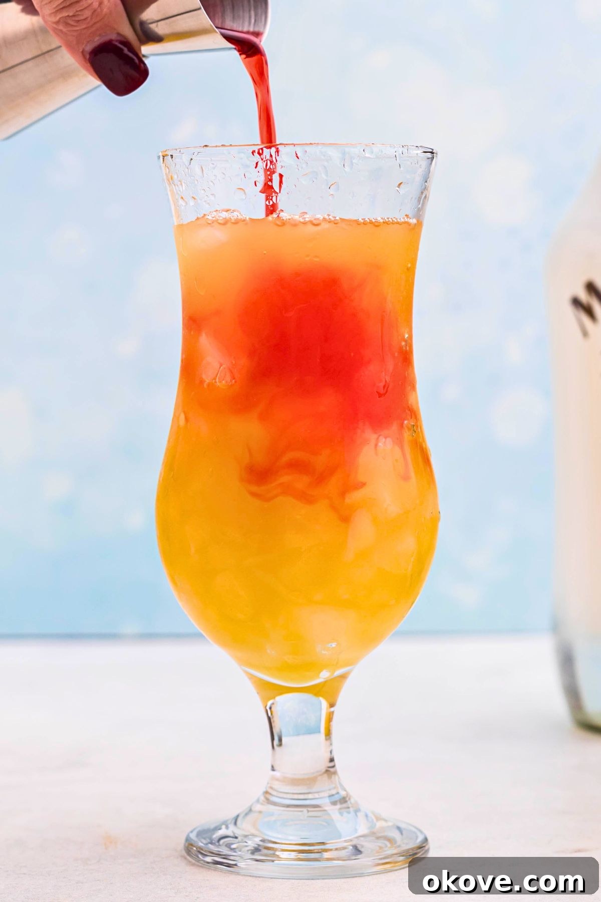 Red grenadine being poured into orange drink  in a hurricane glass. 