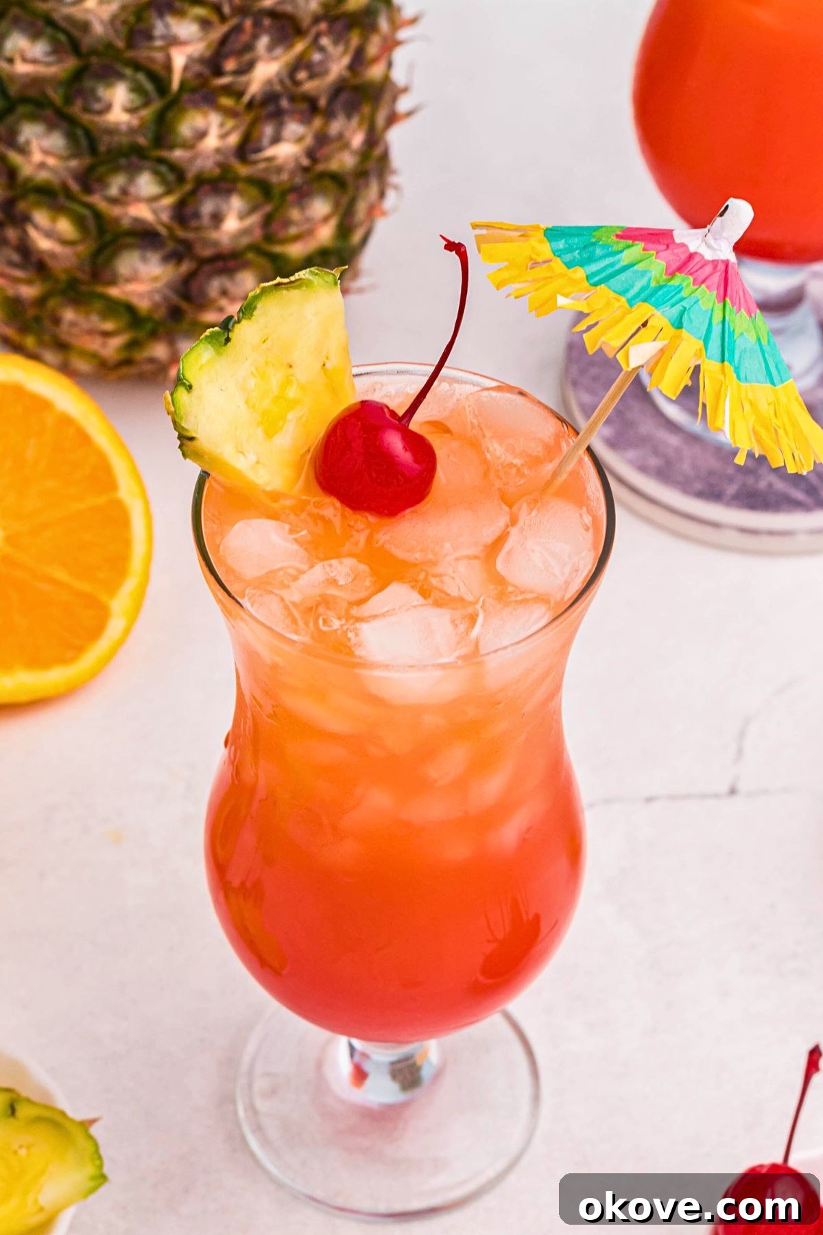 Red and orange drink in a hurricane glass, topped with a cherry, pineapple slice, a nd paper umbrella. 