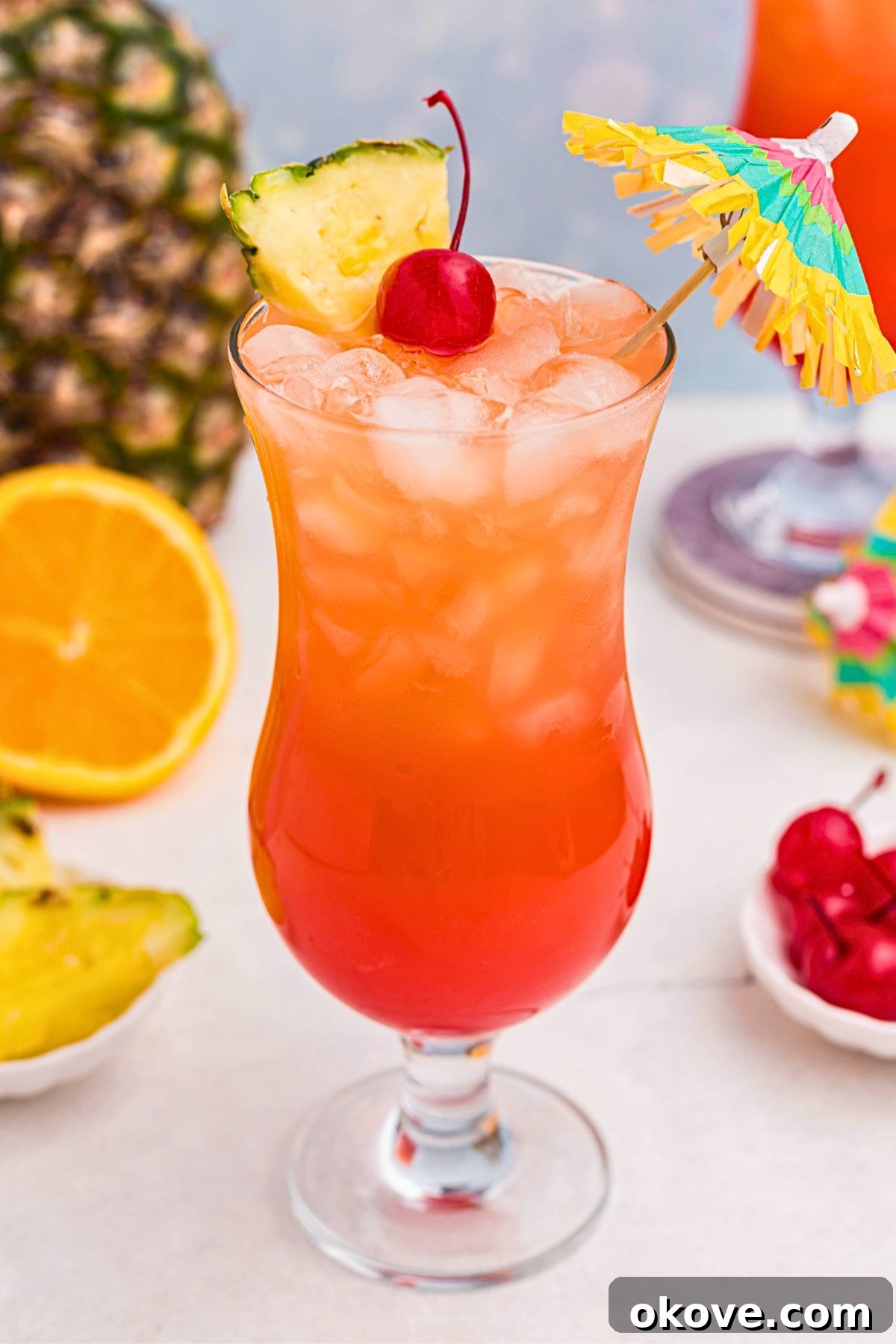 Red and orange drink mixed together, with a paper umbrella, in a hurricane glass with orange slices and cherries on the marble table.