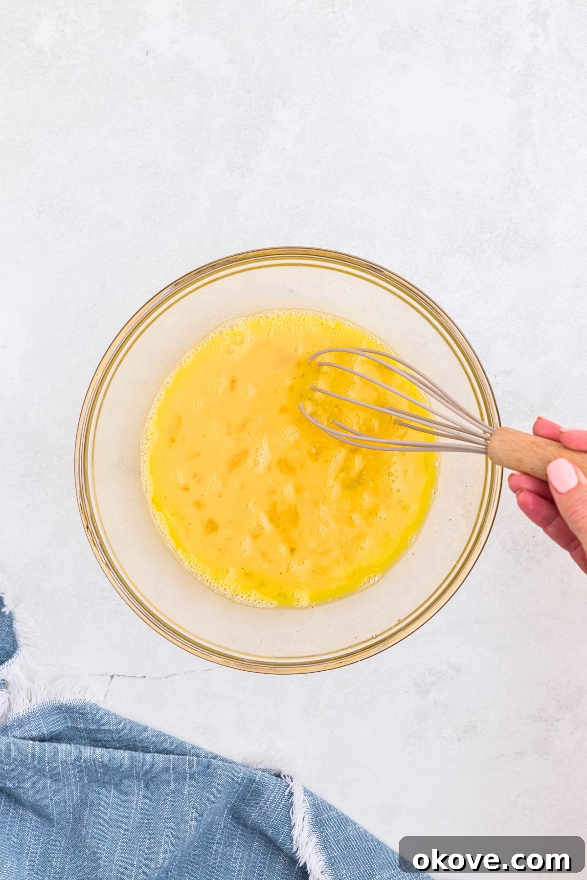 Eggs and seasonings being whisked together with milk in a clear glass bowl.