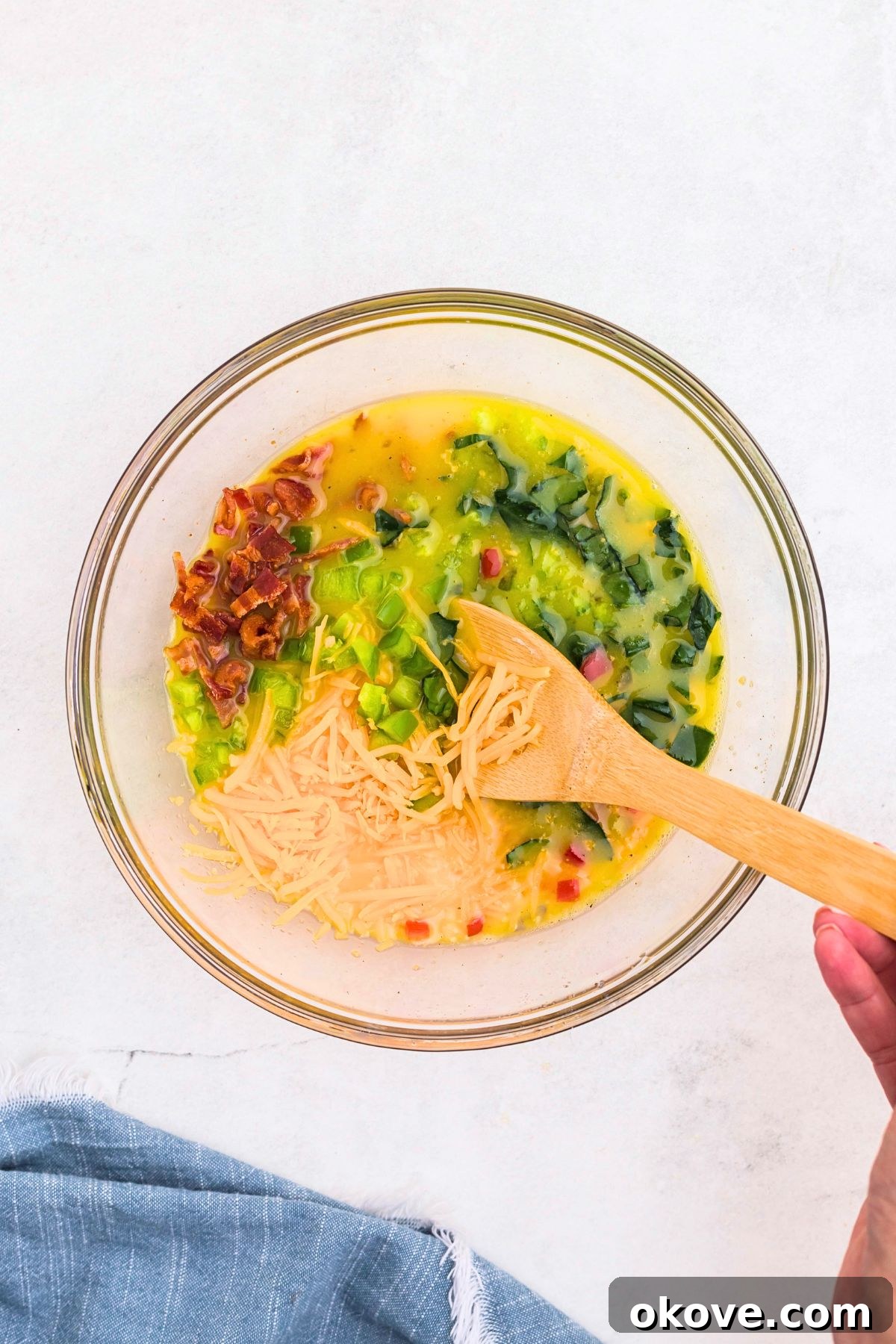 Bell peppers, cheese, and other ingredients being mixed with whisked eggs in clear glass bowl.