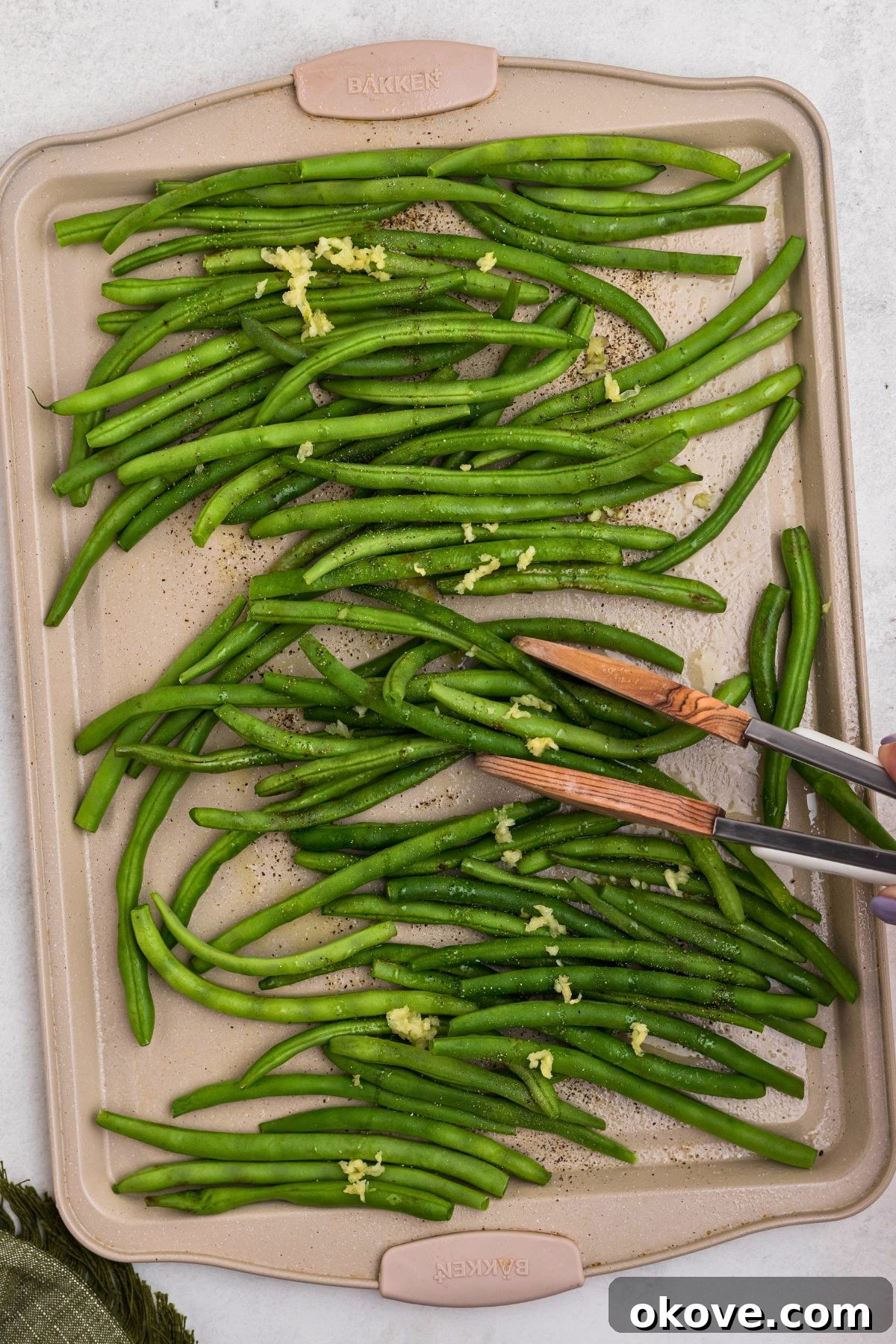 Crispy Roasted Green Beans 5 Fresh green beans evenly spread on a baking sheet, being tossed with olive oil, salt, and pepper using tongs.