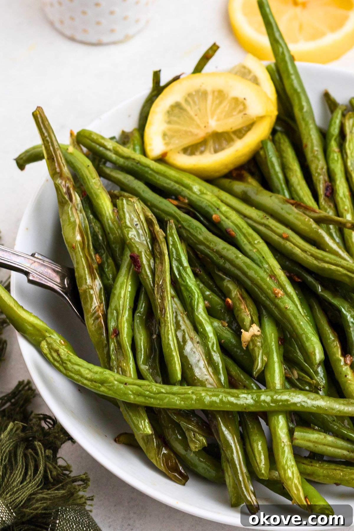 Crispy Roasted Green Beans 8 Fresh green beans arranged in a white bowl, with a serving spoon lifting some beans, accompanied by fresh lemon slices.