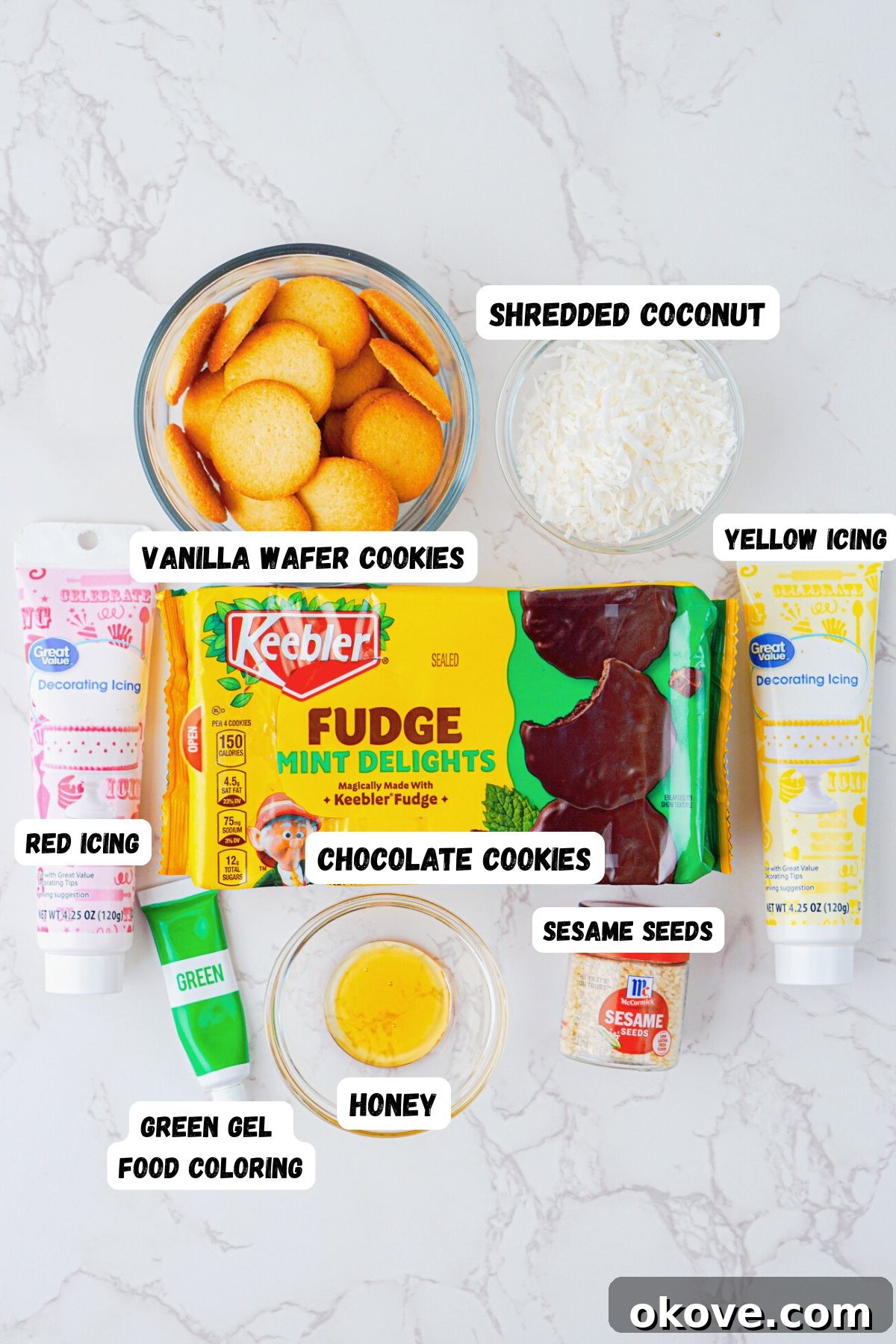 Dessert Burgers 3 A beautiful flat lay of all ingredients laid out on a white marble table: vanilla wafers, fudge mint cookies, shredded coconut, food coloring, yellow and red icing, sesame seeds, and honey, ready to make hamburger cookies.