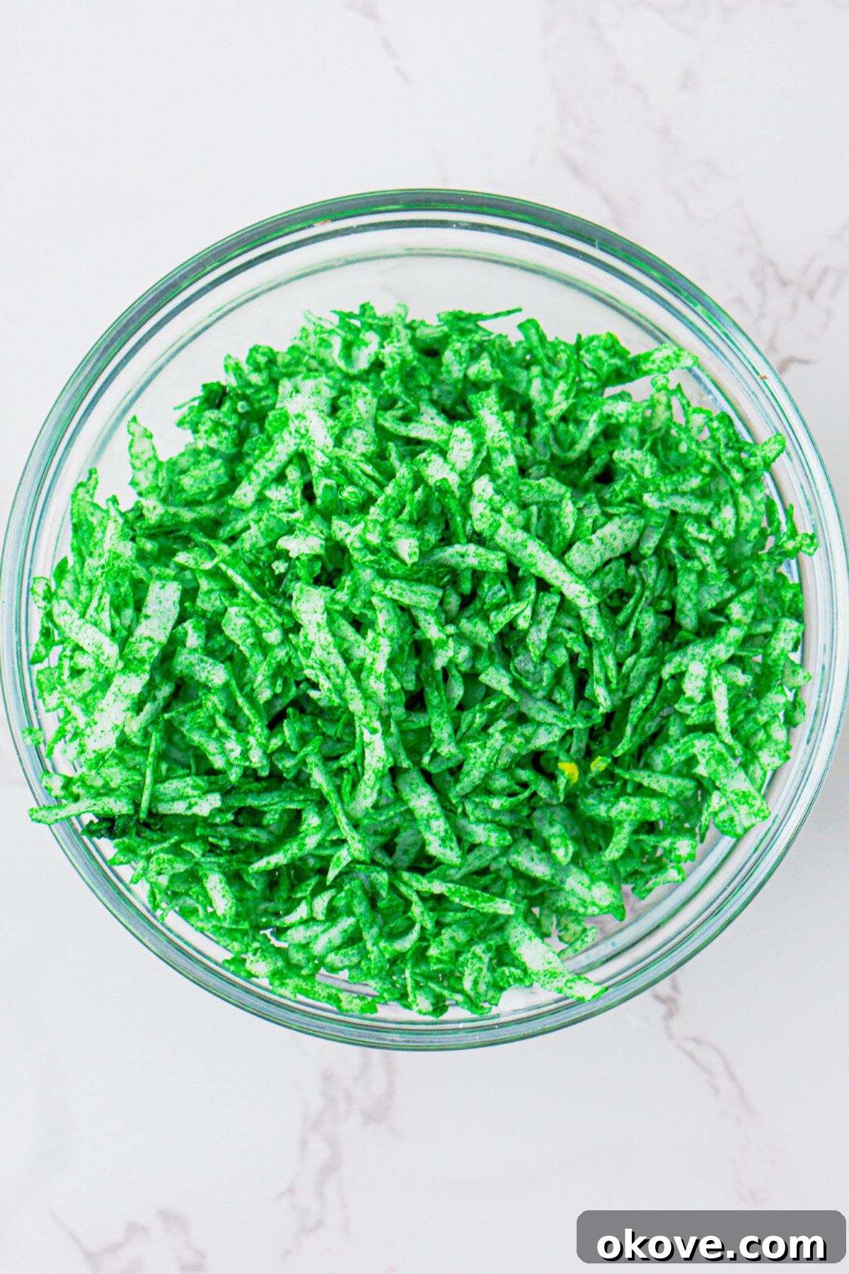 Dessert Burgers 4 Shredded coconut with green food coloring added, in a clear glass bowl, being stirred to create the 'lettuce' layer for the hamburger cookies.