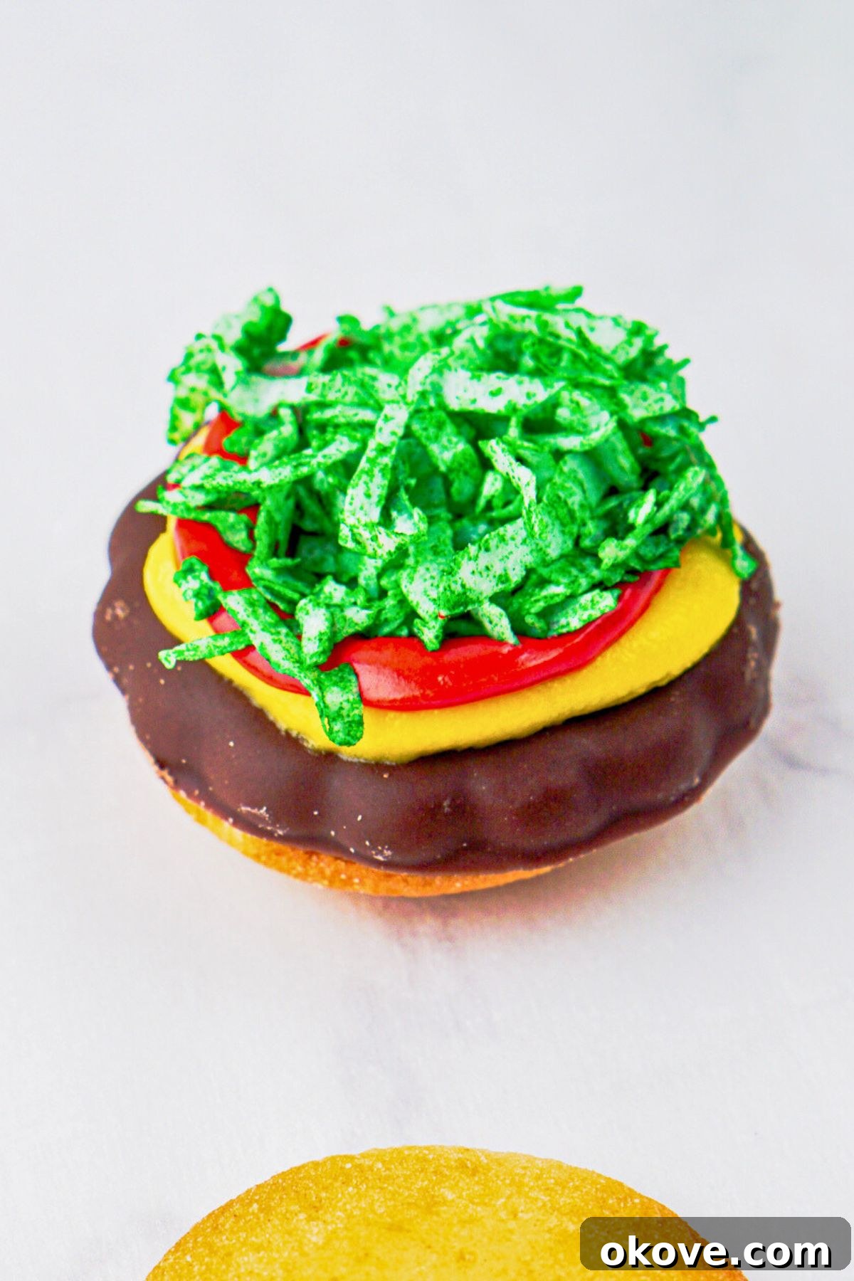 Dessert Burgers 6 Assembled hamburger cookie showing the vanilla wafer bottom bun, chocolate patty with red and yellow icing, and a generous sprinkle of green shredded coconut on a white marble board.