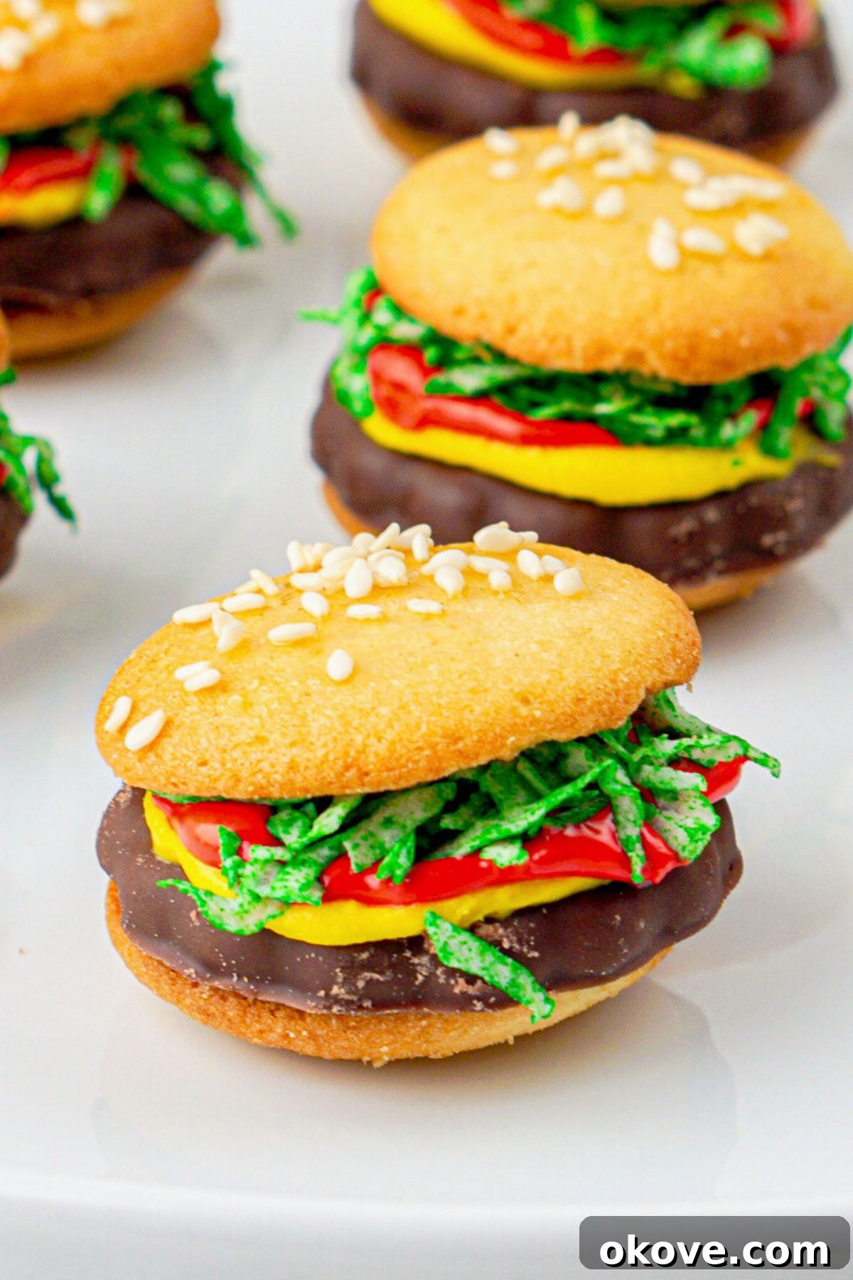 Dessert Burgers 8 A close-up shot of a plate of perfectly assembled vanilla wafer cookies resembling hamburgers, complete with frosting, shredded coconut 'lettuce,' chocolate cookie 'patties,' and sesame seeds on top. These no-bake treats are ready to impress.