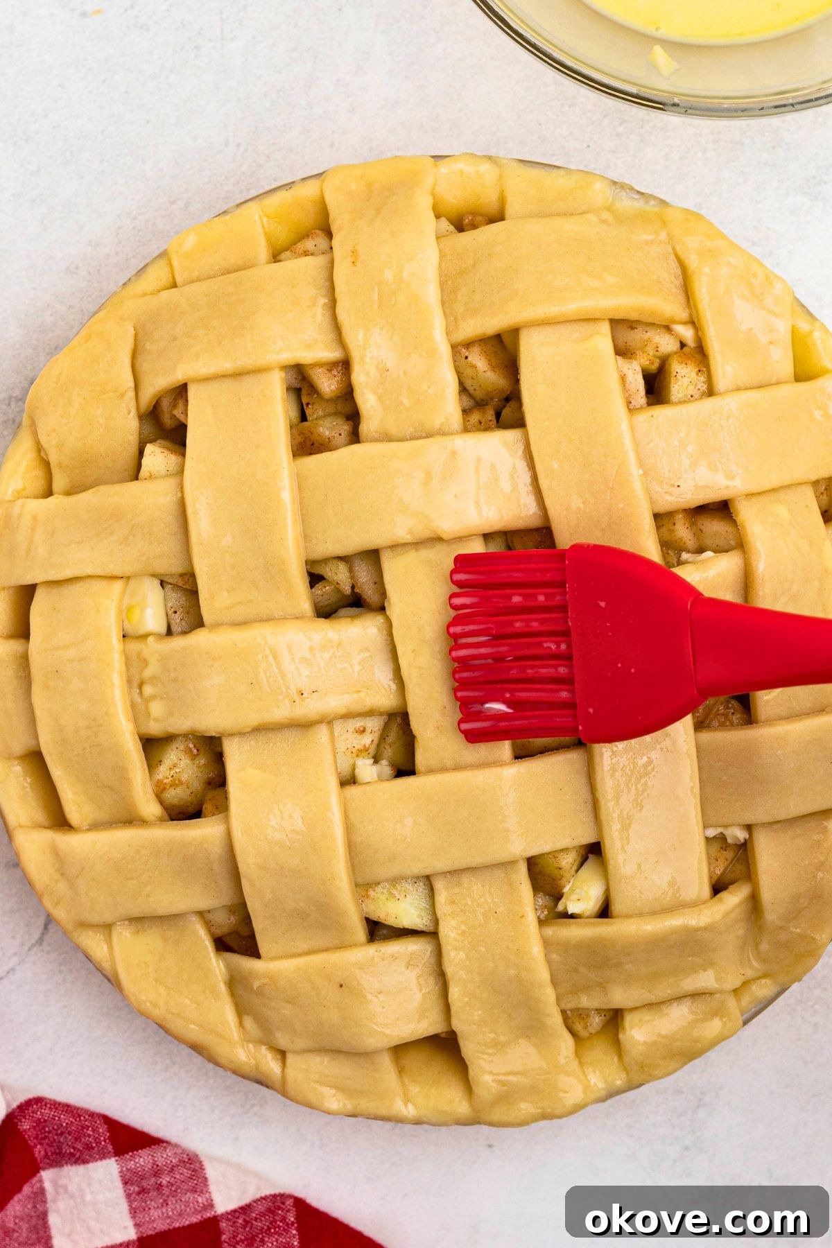 Uncooked apple pie with egg wash being brushed on top with a red pastry brush. 