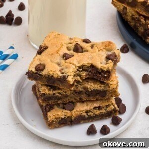 Cookie bars stacked on a small white plate in front of a tall glass of milk, with chocolate chips scattered on the table.