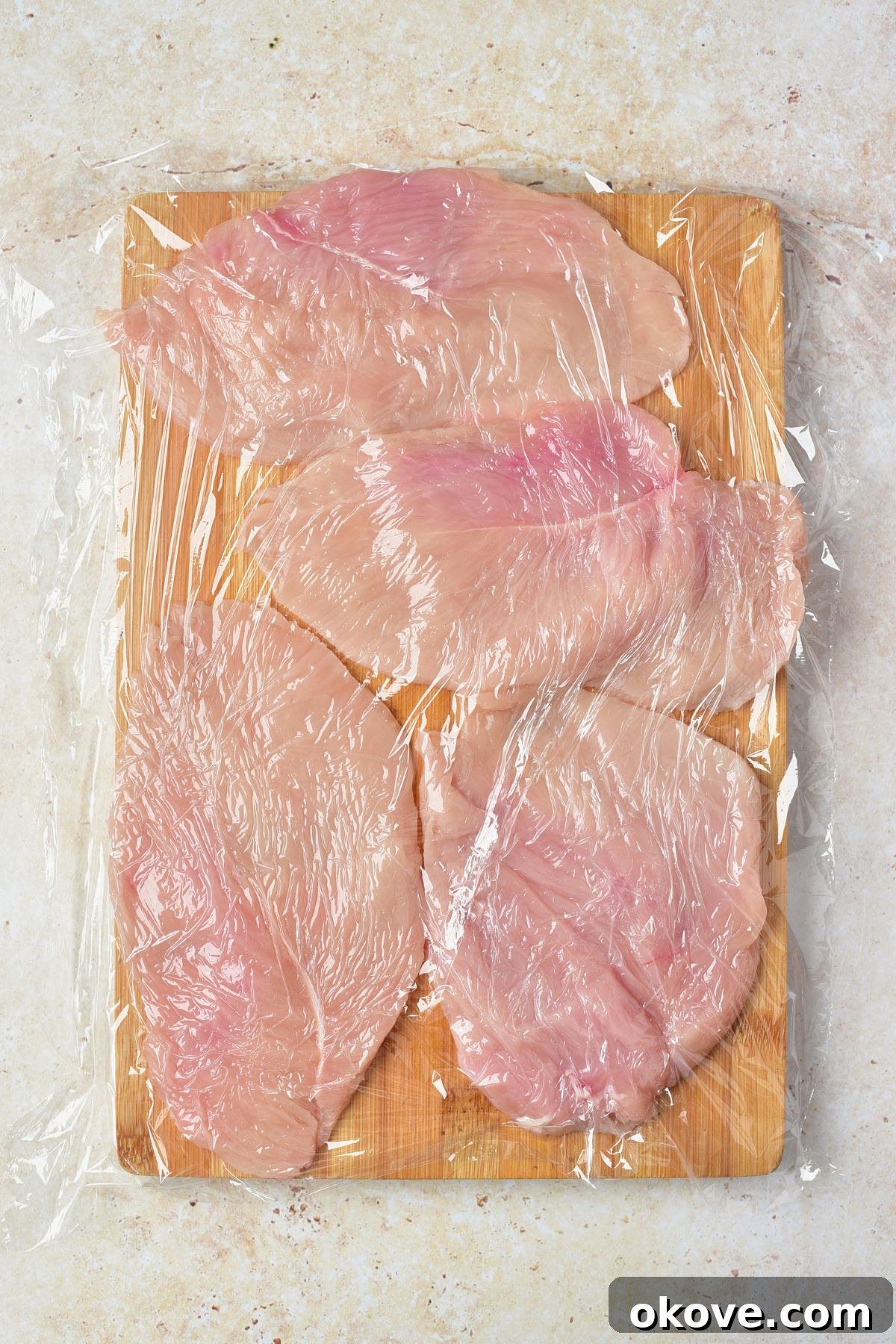 Chicken breasts flattened into even thickness, covered with plastic wrap, on a wooden cutting board.