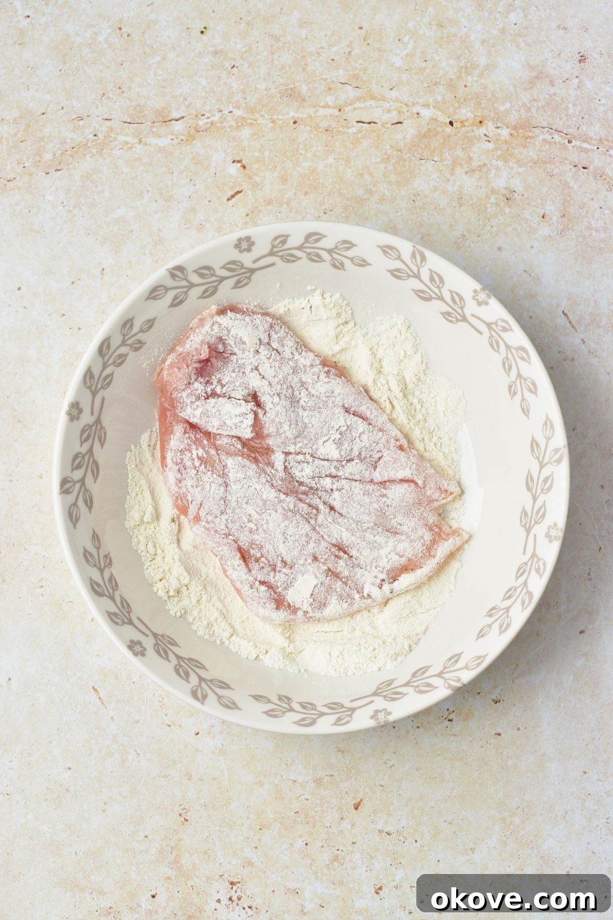 Uncooked chicken breasts coated in flour, in a shallow bowl.