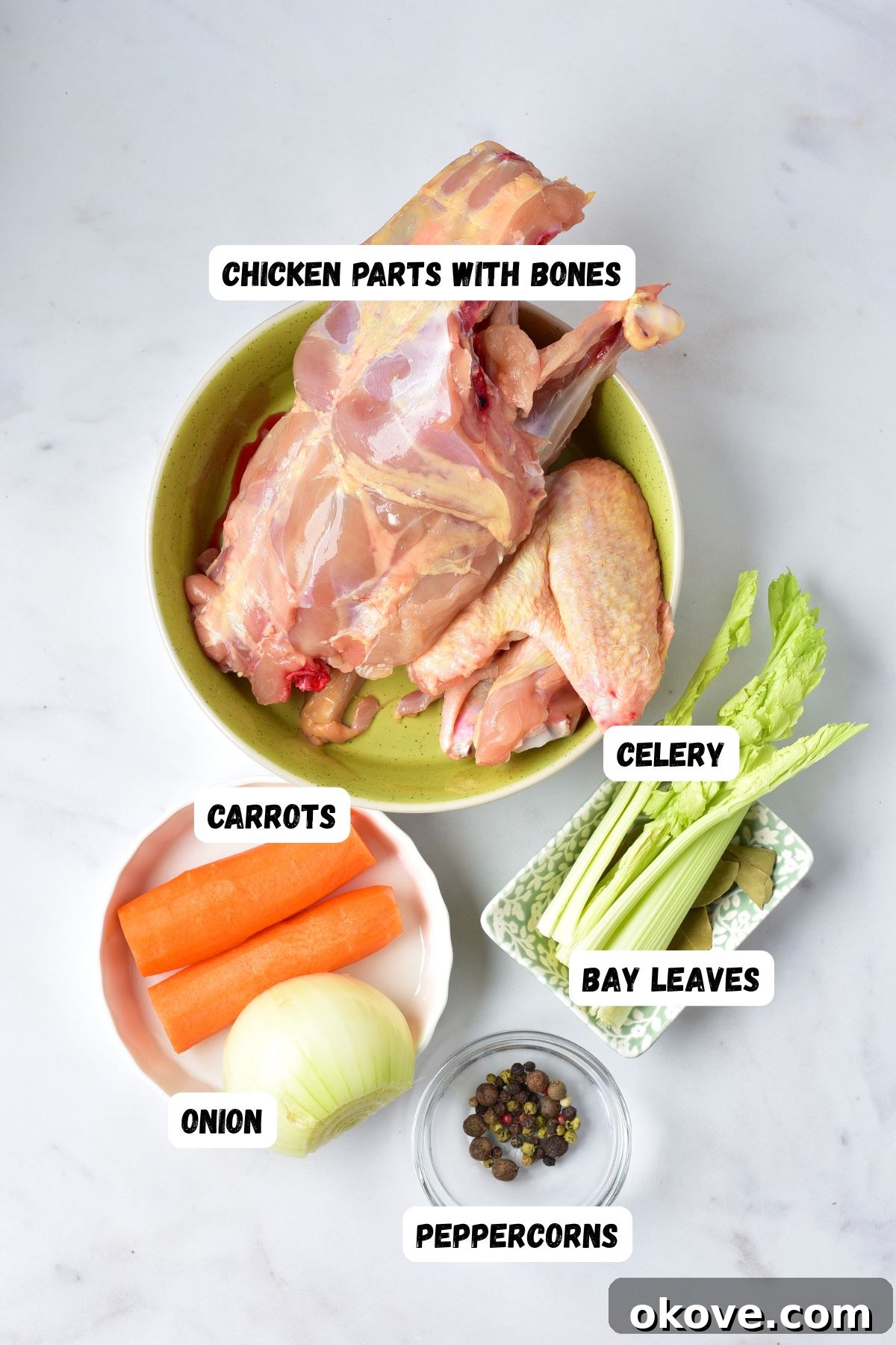 Ingredients needed to make chicken stock measured into small bowls on a white marble table, including chicken bones, carrots, onions, celery, bay leaves, allspice, and peppercorns.