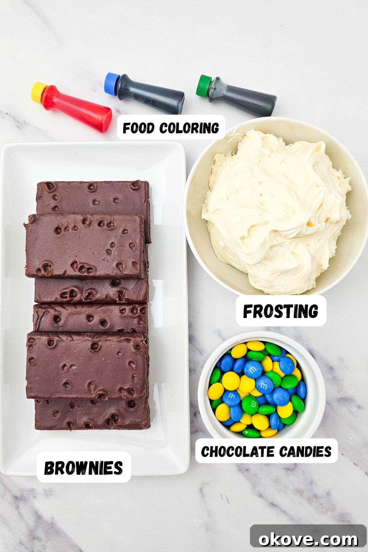 Brownies on a plate, white frosting in bowls, and small blue, green, and yellow M&M candies in separate bowls, with bottles of food coloring, all arranged on a marble table.