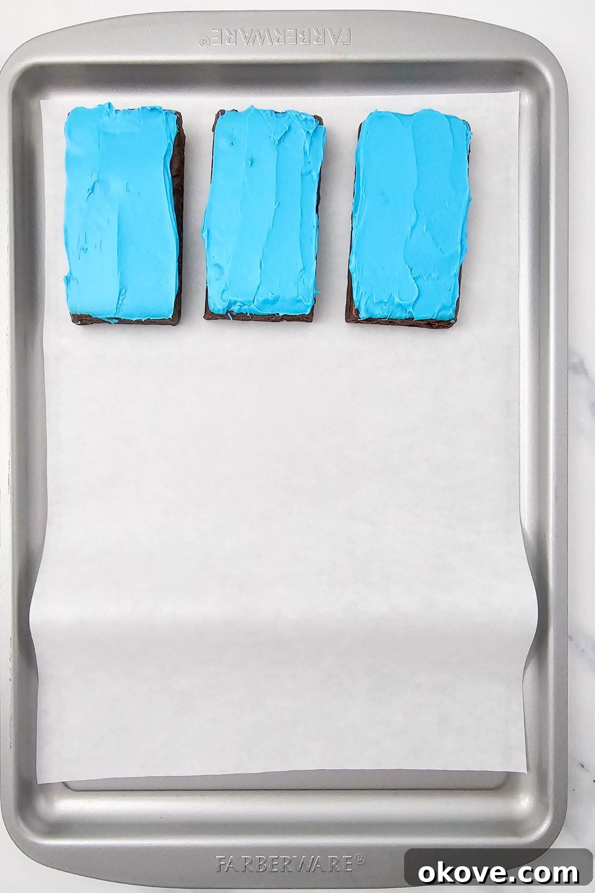 Brownies being frosted with colorful frosting and placed on a baking sheet, lined with parchment paper.