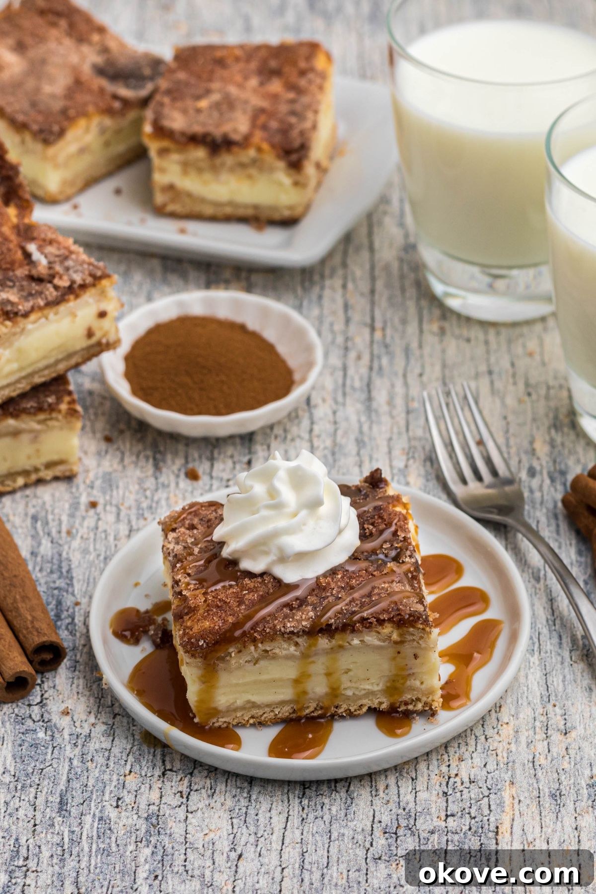 Cinnamon Sugar Crust Cheesecake 2 Slice of churro cheesecake bar on a small white plate, drizzled with caramel syrup and topped with whipped cream.
