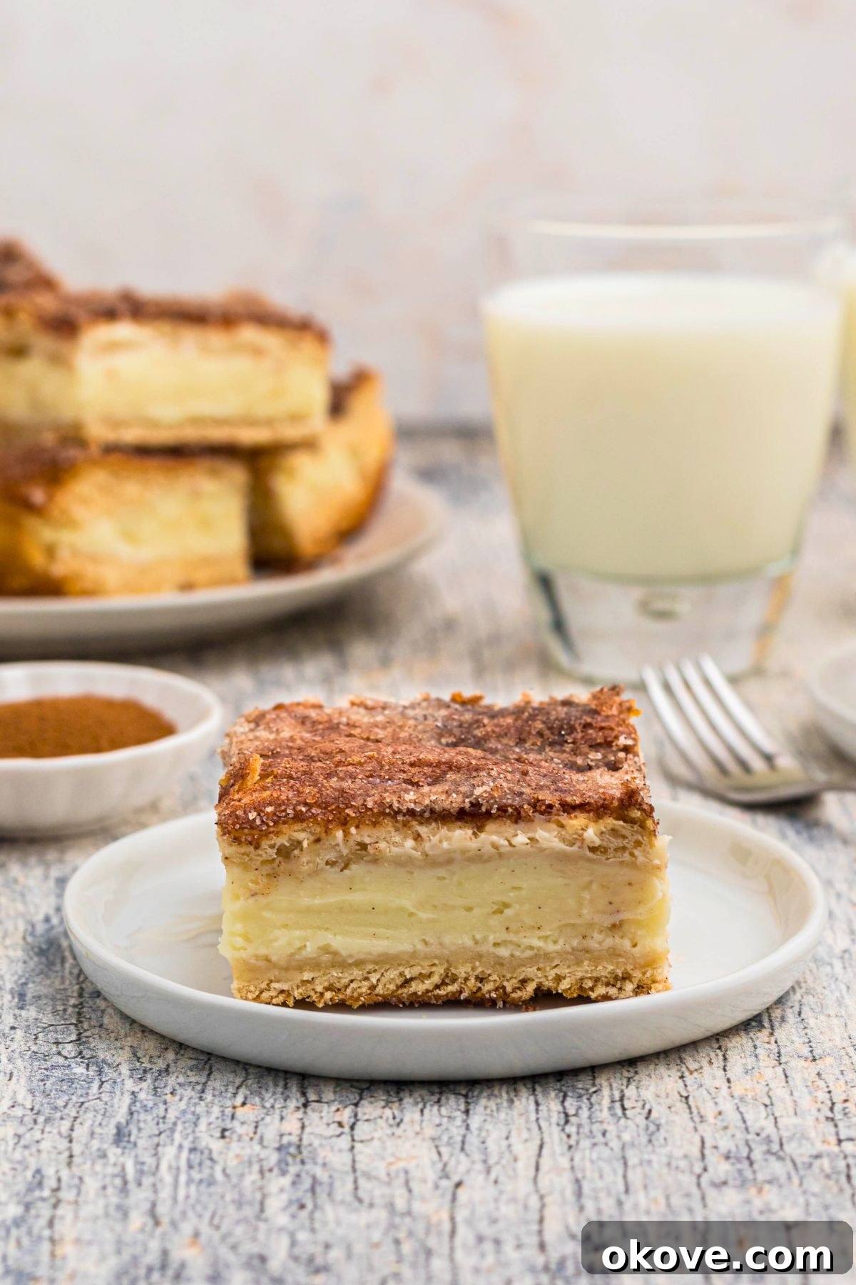 Cinnamon Sugar Crust Cheesecake 12 Slice of churro cheesecake served on a small white plate in front of glasses of milk, drizzled with caramel.