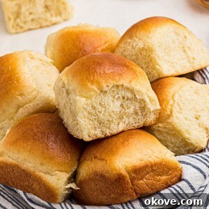 Golden, perfectly baked dinner rolls nestled in a small bowl, lined with a blue and white linen, resting on a pristine white marble table.
