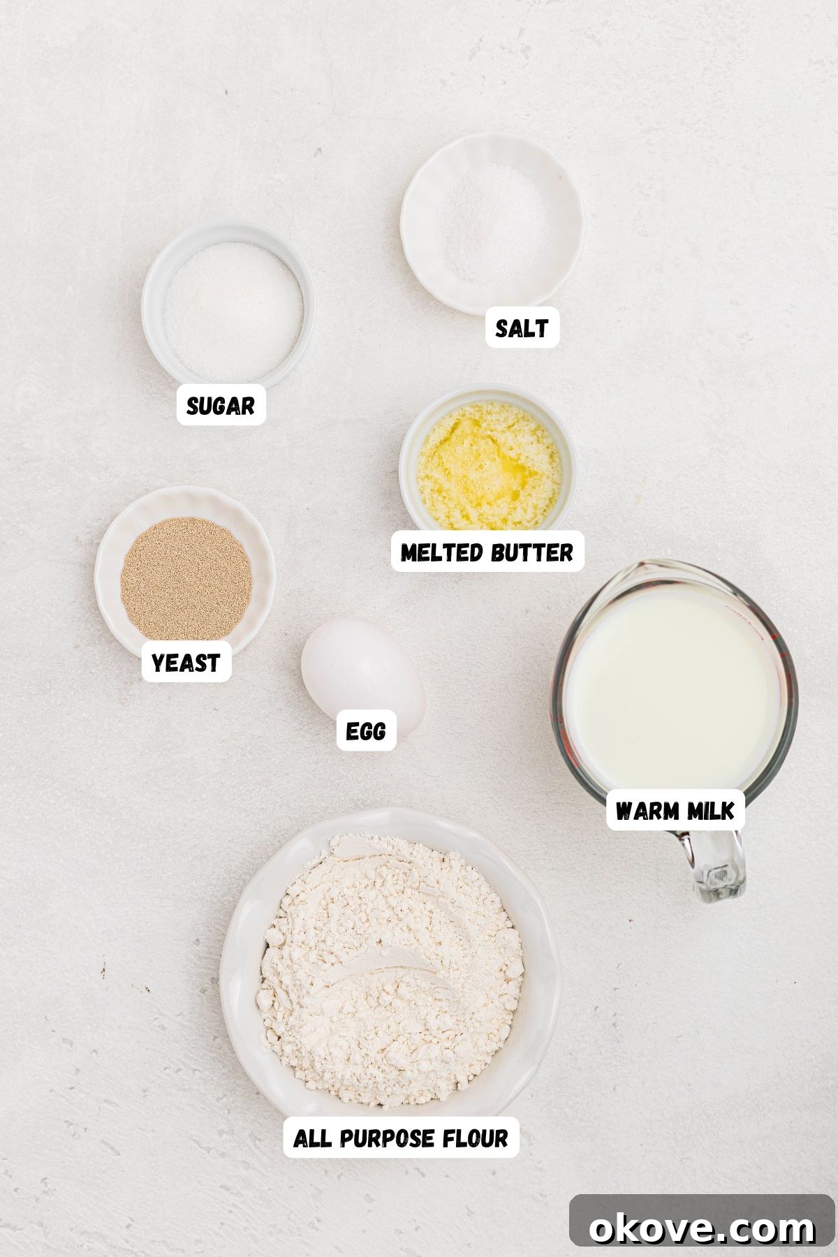 Warm Golden Dinner Rolls 3 All the essential ingredients meticulously measured into small, clear bowls, neatly arranged on a white marble table, ready for baking homemade dinner rolls. Ingredients include yeast, milk, sugar, egg, butter, flour, and salt.