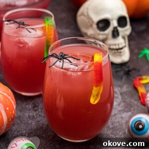 Glass of Halloween Punch in with gummy worms hanging over the side of the glass, in front of plastic skull and spiders.
