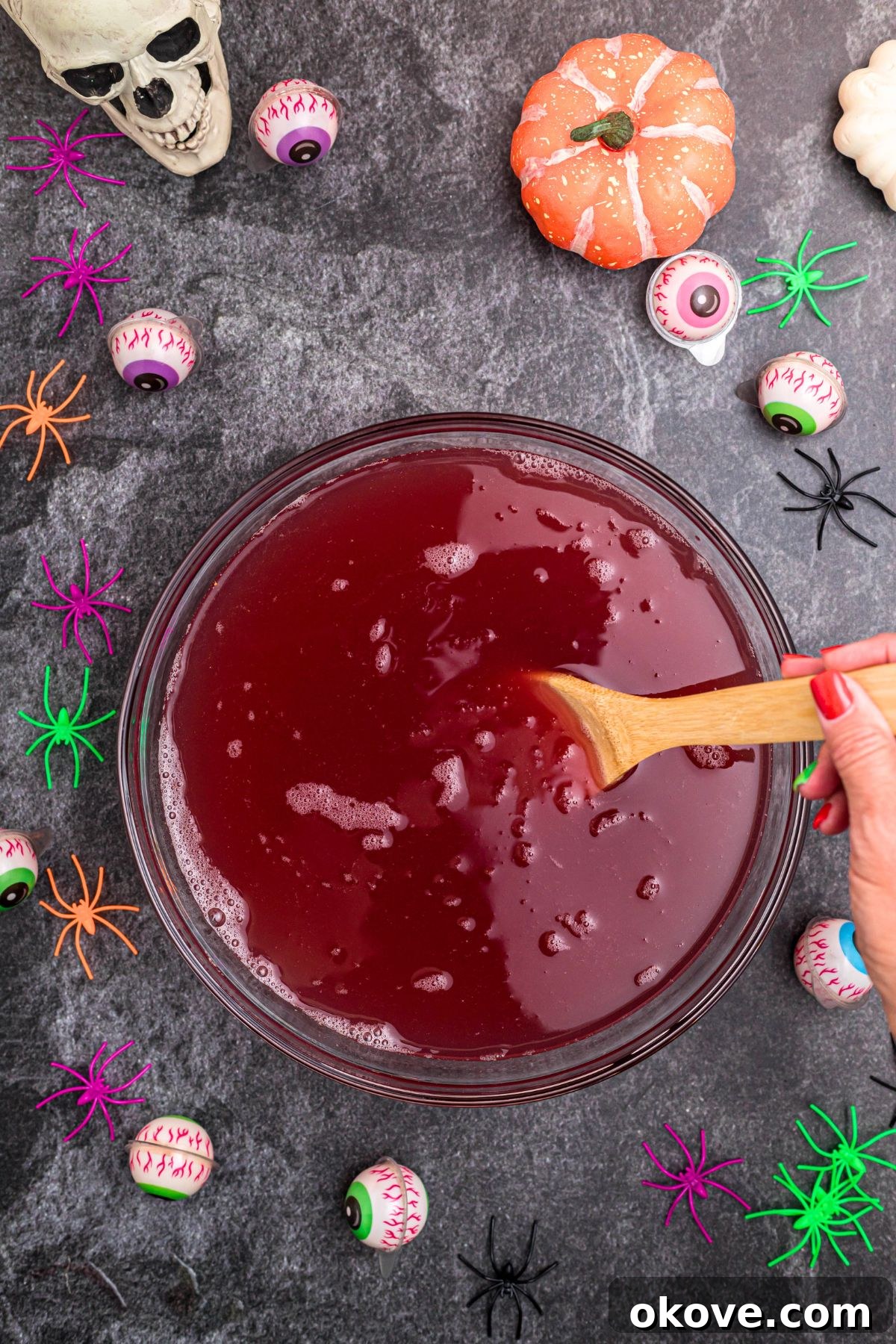 Halloween punch being gently stirred with a wooden spoon in a glass bowl, surrounded by plastic spiders and candy eyes on a black marble table.
