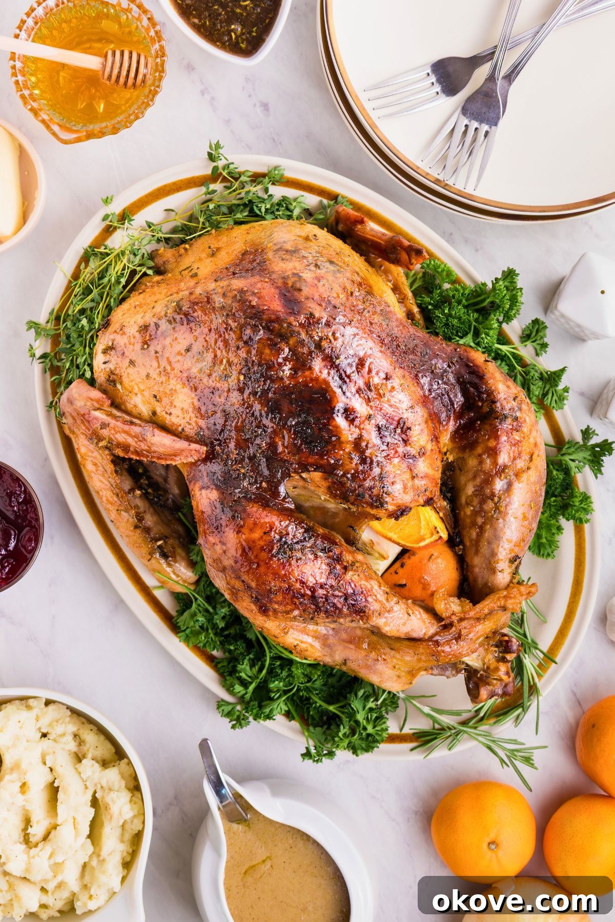 Golden Roasted Turkey Perfection 2 Plated roasted turkey on a serving platter, surrounded by plates, honey, and other food, on a white marble table.