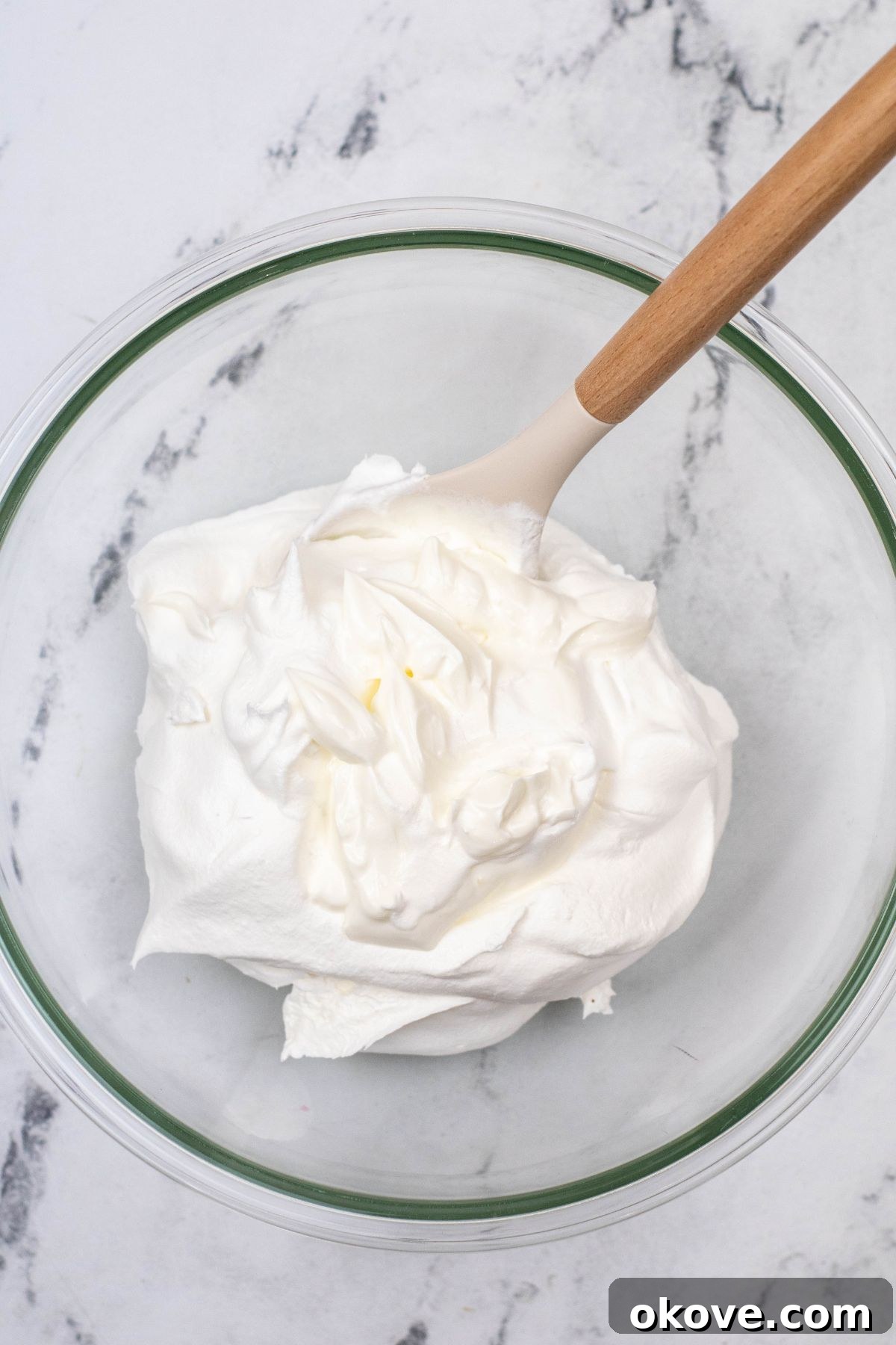 Whipped topping and sour cream being mixed together in a large clear glass bowl until smooth.