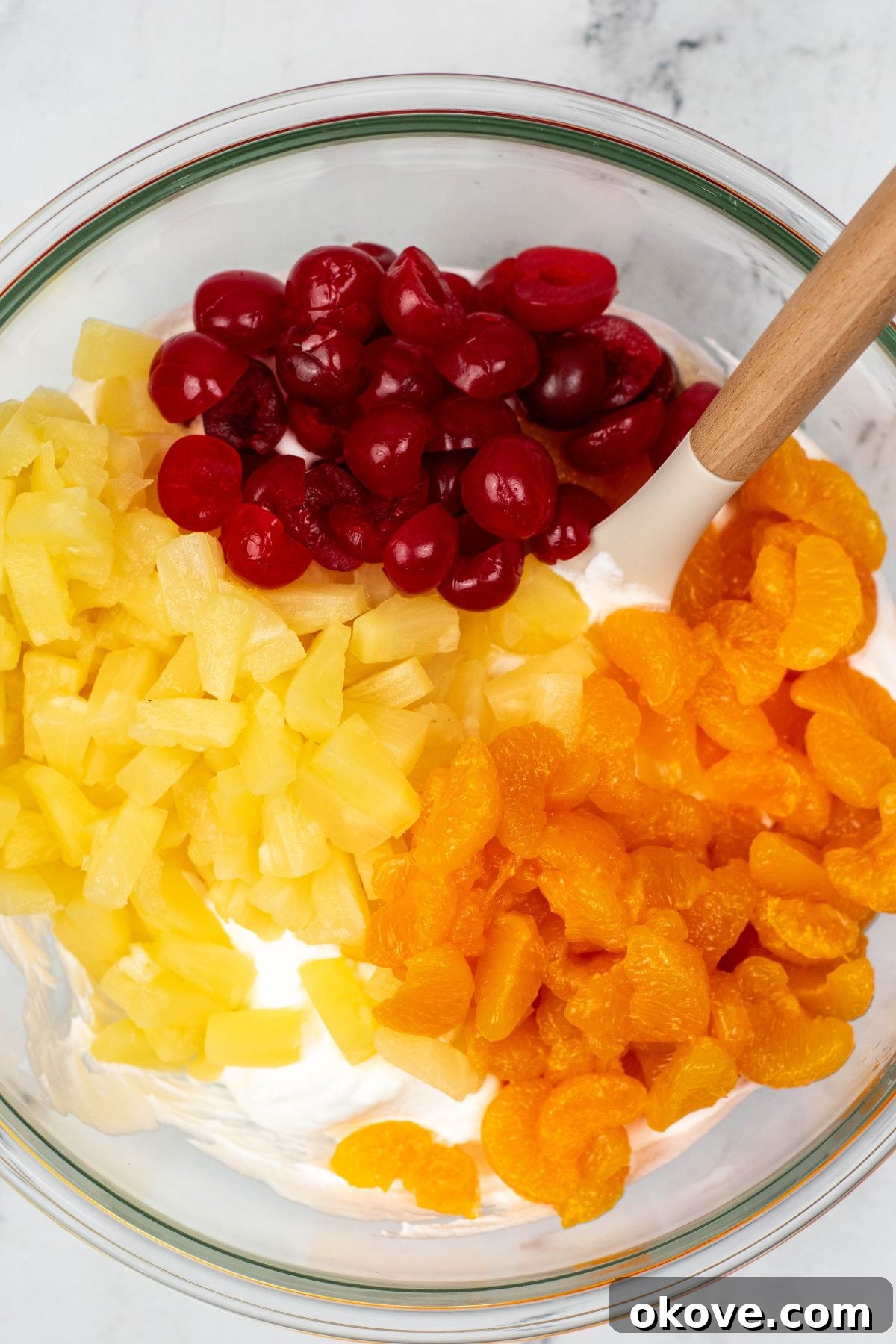 Drained cherries, pineapple, and mandarin oranges being carefully added to the whipped cream mixture in a large glass bowl.