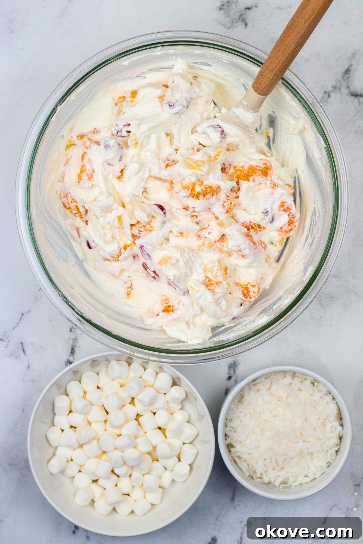 A large clear glass mixing bowl, filled with the fruit and whipped cream mixture, now having mini marshmallows and shredded coconut added and gently folded in.