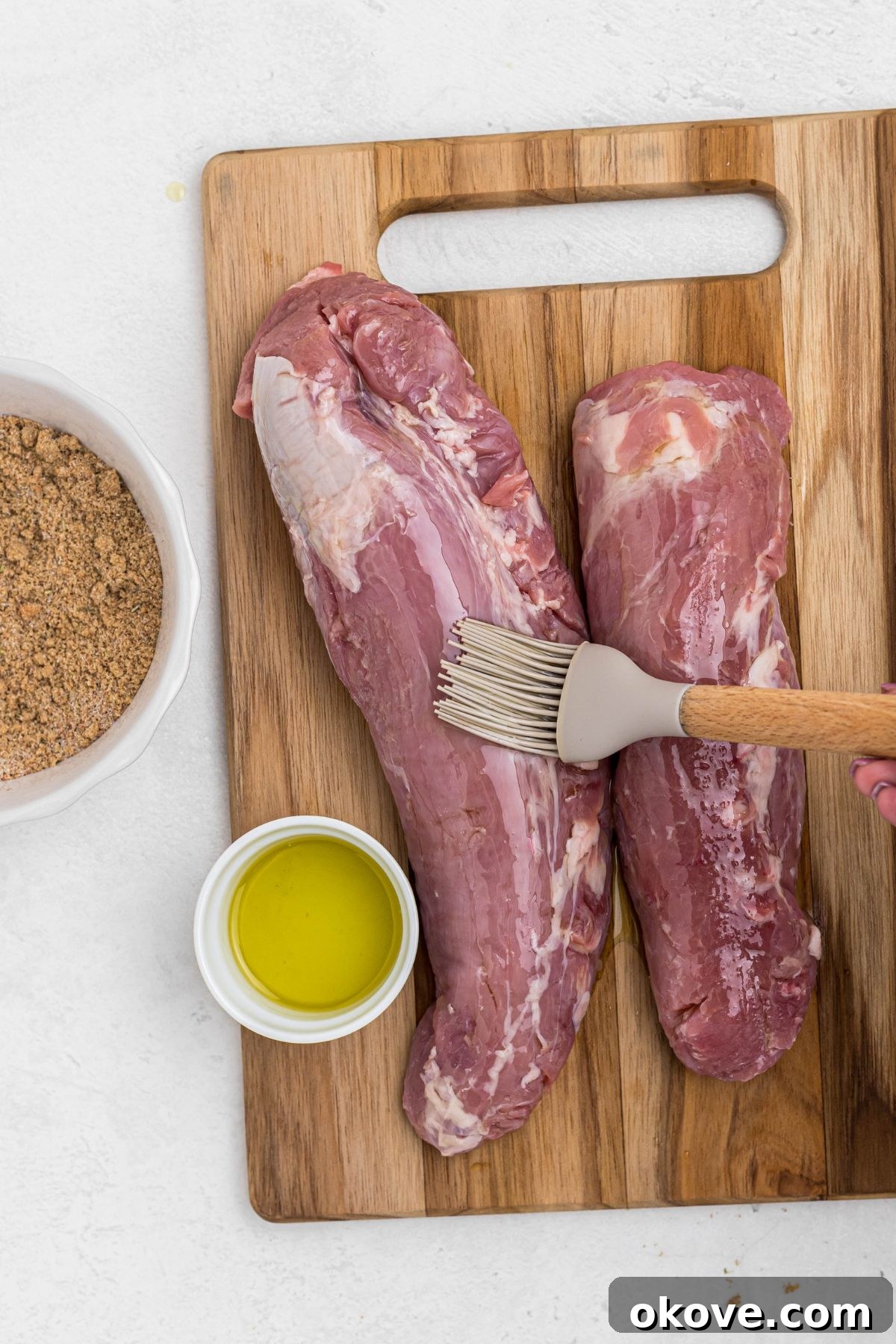 Smoky Pork Tenderloin Perfection 4 Two uncooked pork tenderloins on a wooden cutting board with olive oil being brushed on top, preparing them for the dry rub.