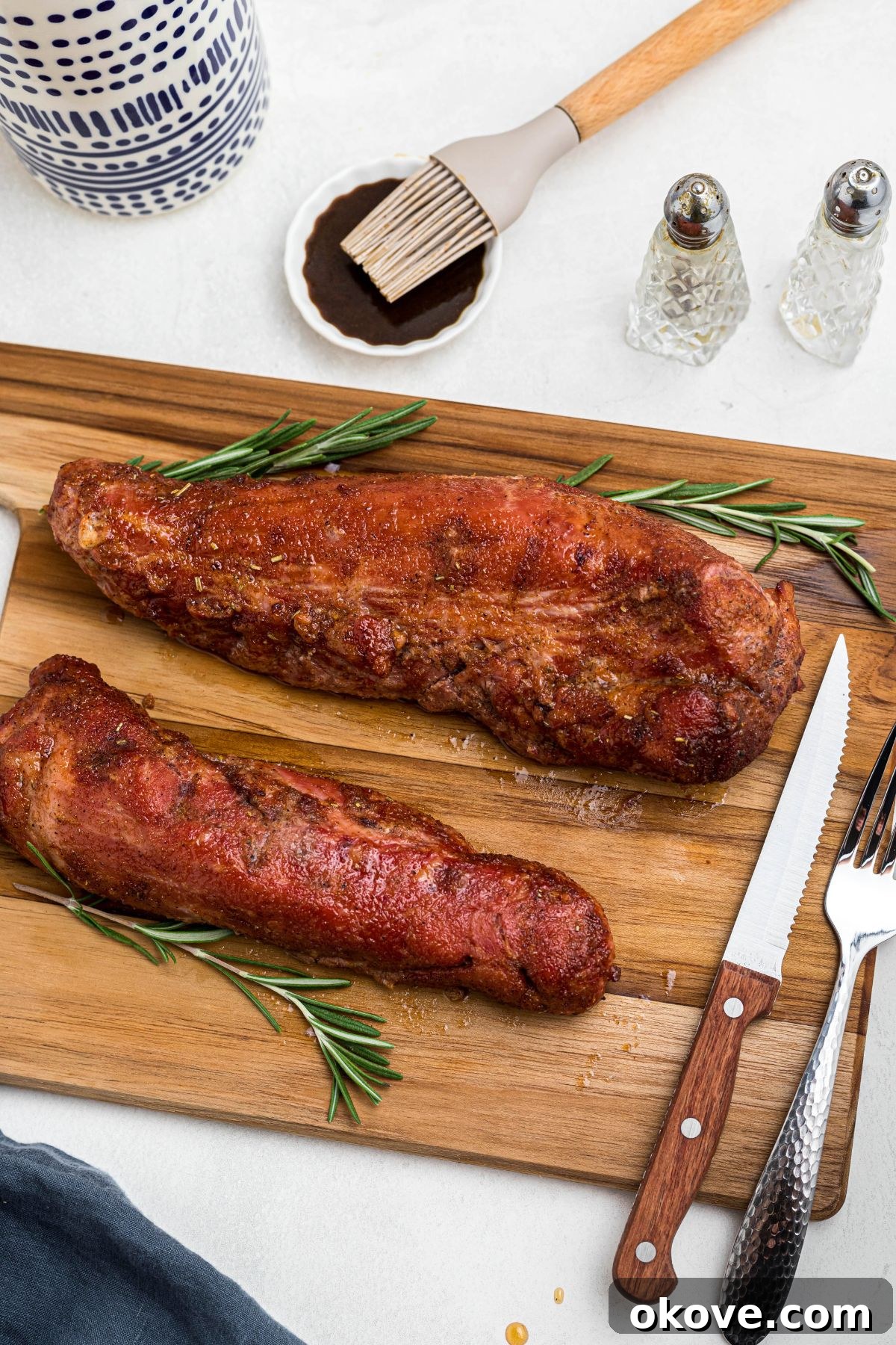 Smoky Pork Tenderloin Perfection 9 Smoked tenderloins resting on a wooden cutting board, glistening with glaze, with additional sauce being brushed on the side for serving. The meat is ready to be sliced.