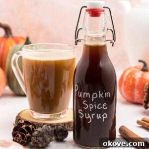 Bottle of pumpkin spice syrup on a small wooden coaster next to a cup of coffee, on a marble table next to a cup of coffee.