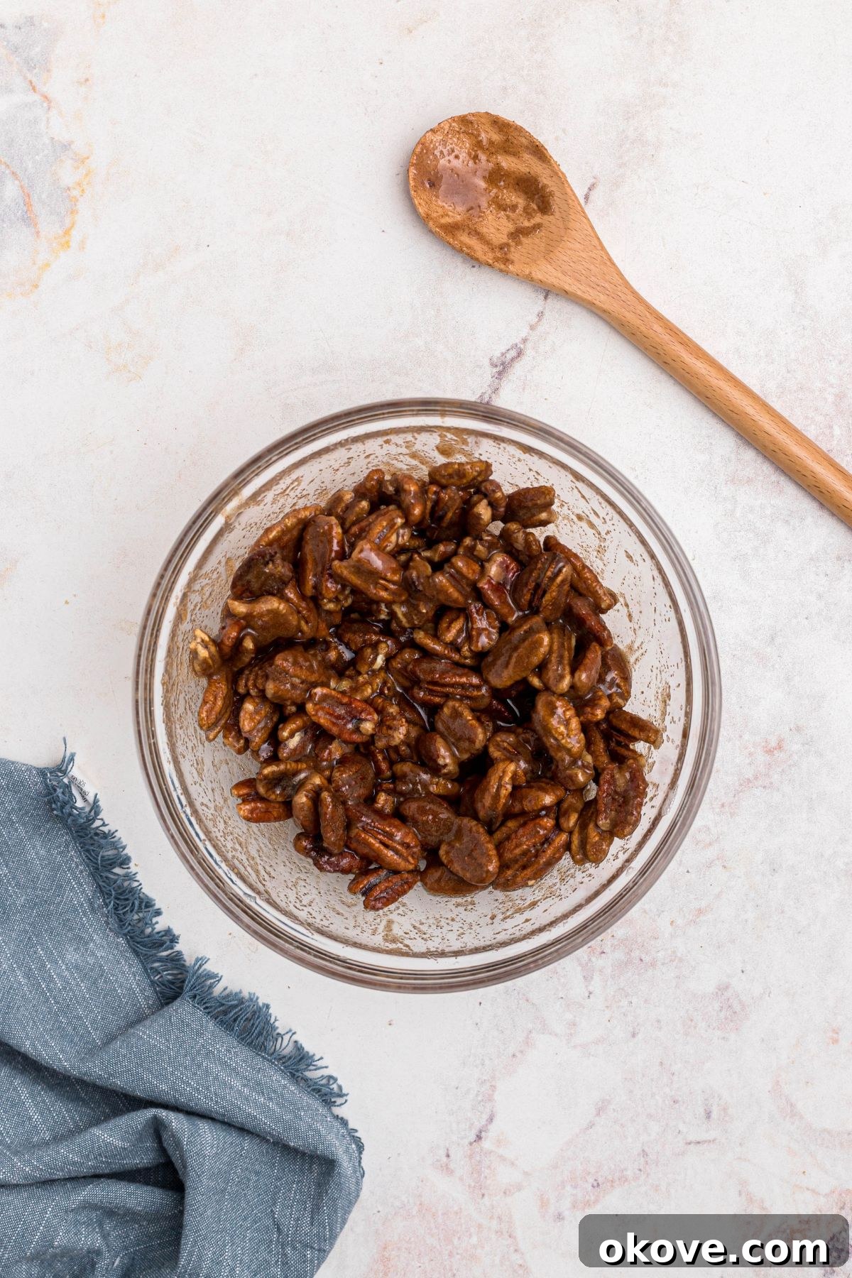 Sweet Pecan Crunch 7 Sugars and cinnamon being tossed with egg-white coated pecans in a clear glass bowl on a marble table.
